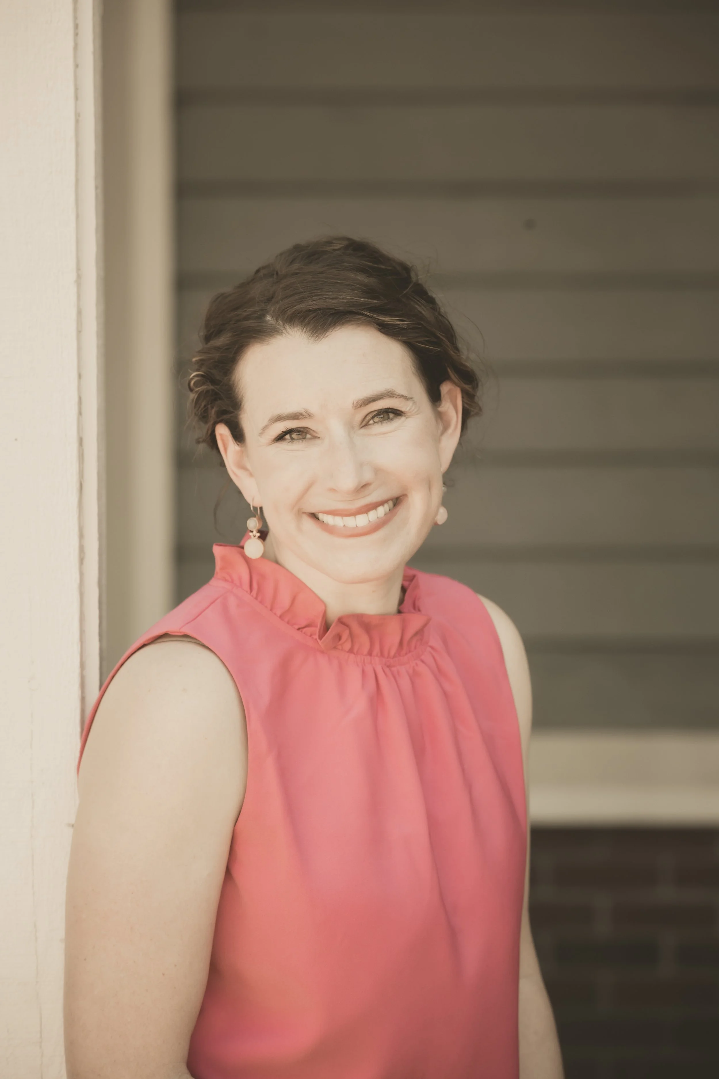 Close-up of a smiling woman with short brown hair, wearing a pink sleeveless top and earrings, standing outdoors against a background of horizontal siding and a wall.