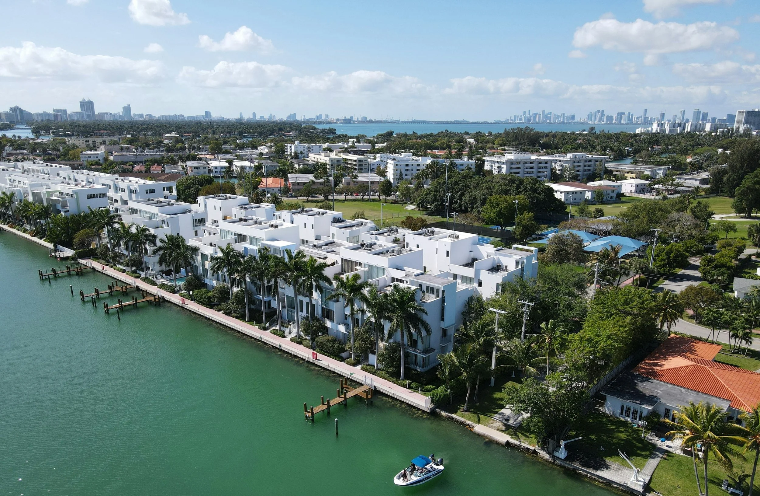 Aerial view of white modern waterfront homes with docks along a green canal, lush trees, and a city skyline in the background under a partly cloudy sky.