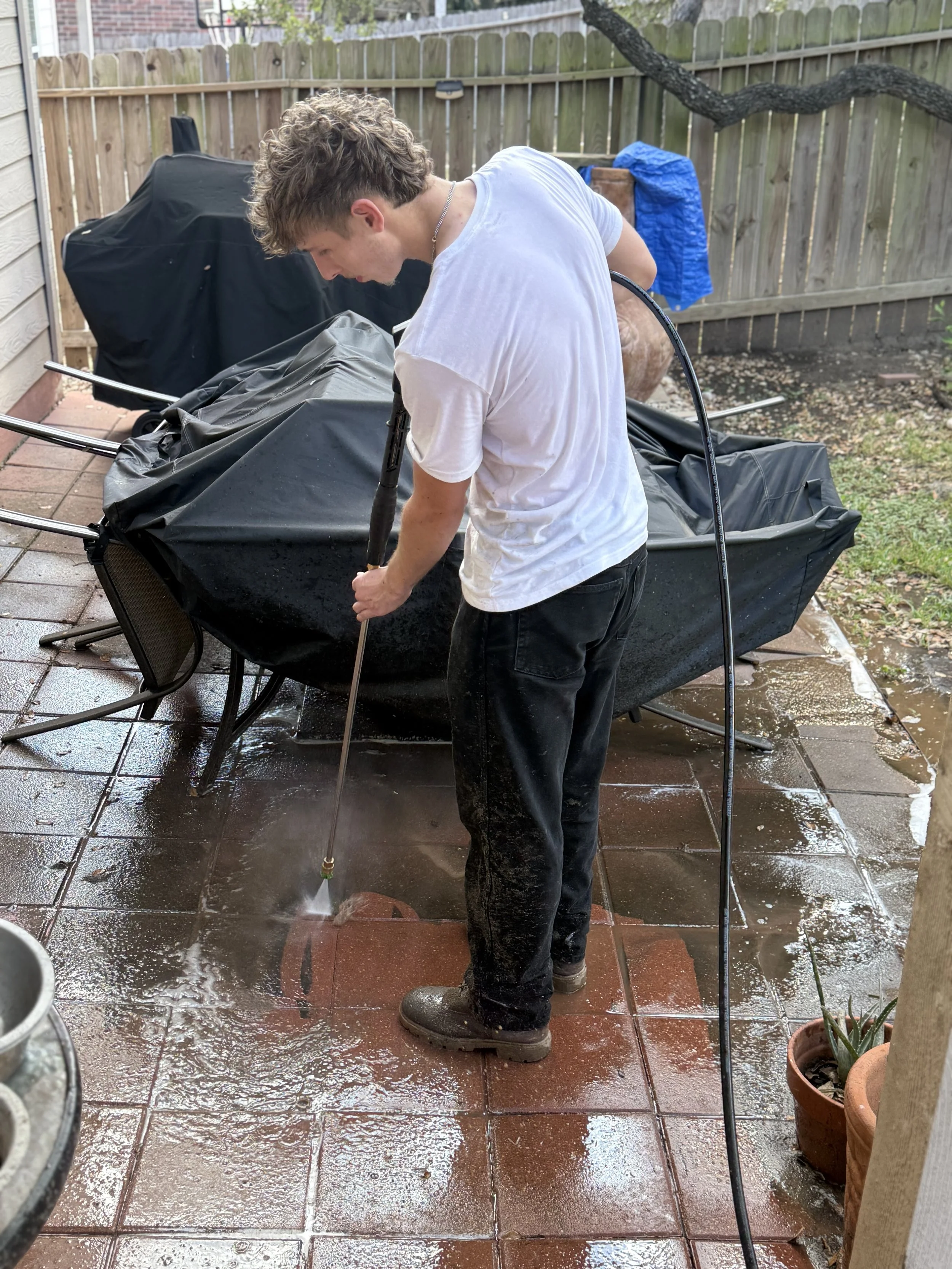 A young man wearing a white t-shirt, black pants, and boots is pressure washing a tiled patio. He is bent over, holding a pressure washer wand, and water is splashing on the wet surface. There is a wooden fence, outdoor furniture covered with black and blue covers, and potted plants in the background.