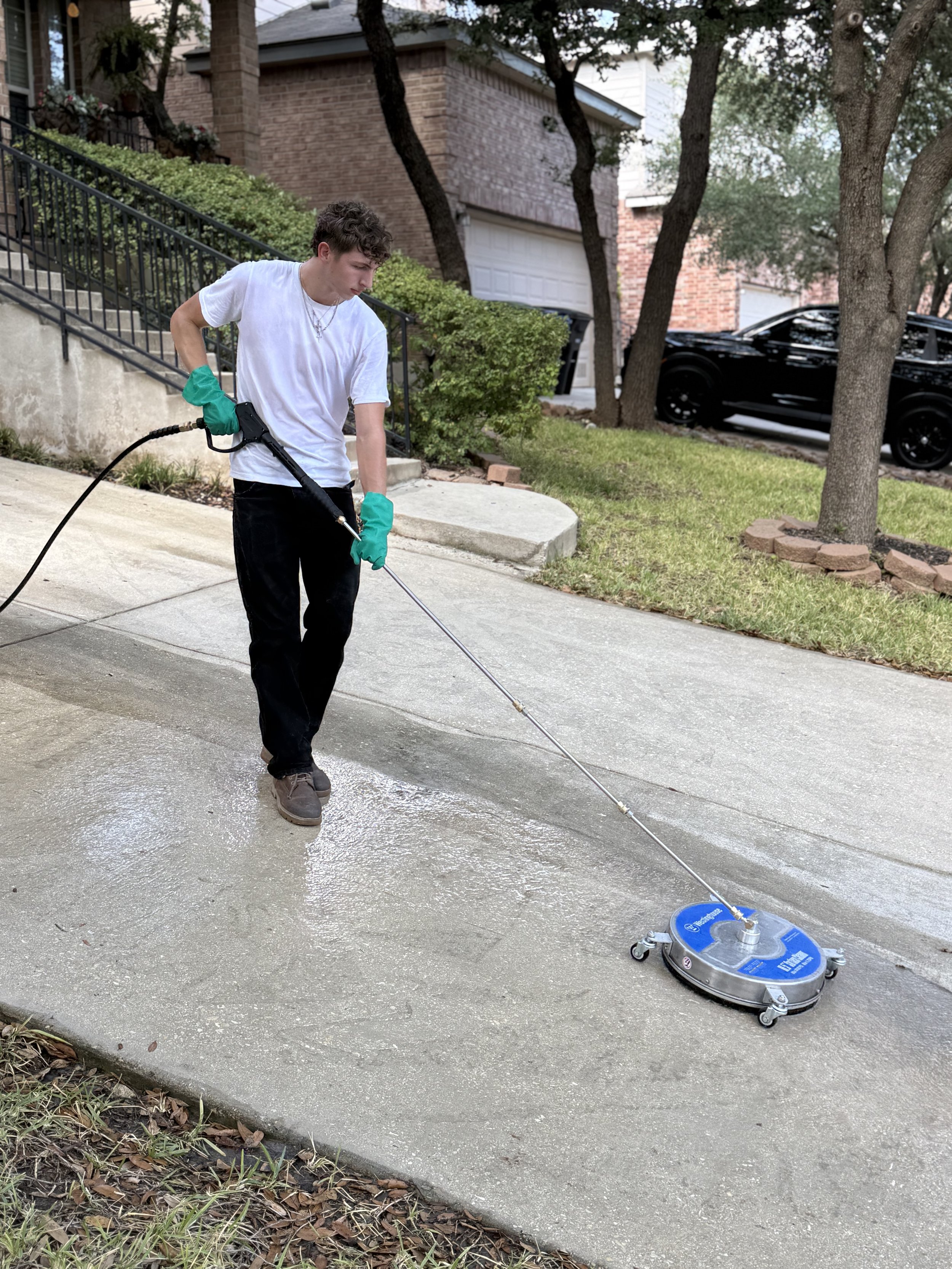 Person wearing white t-shirt, black pants, and green gloves using a floor cleaning machine on a concrete sidewalk in a residential neighborhood with trees, shrubs, and brick houses.