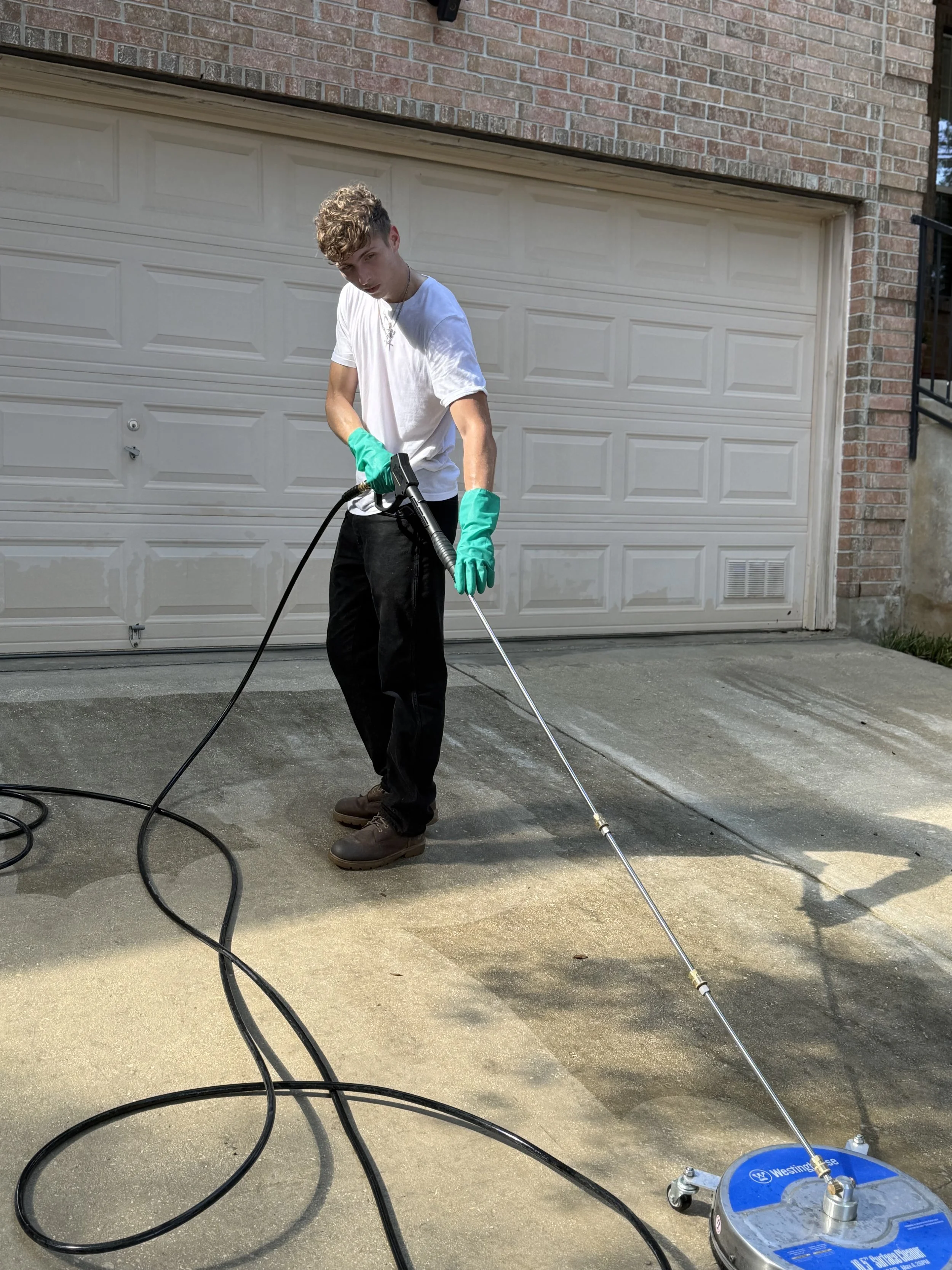 A young man wearing a white t-shirt, black pants, and green gloves is power washing a concrete driveway in front of a brick house with a white garage door.