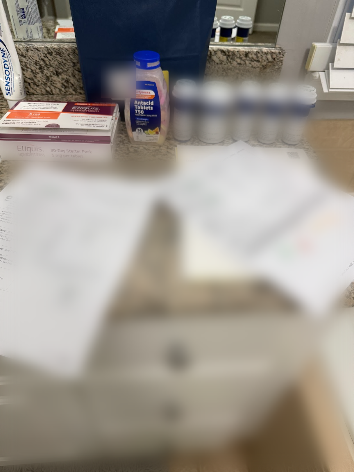 A bathroom counter covered with prescription bottles, an Eliquis 30-day starter pack, antacid tablets and several printed pages of a hospital medication list; names and labels are blurred for privacy.