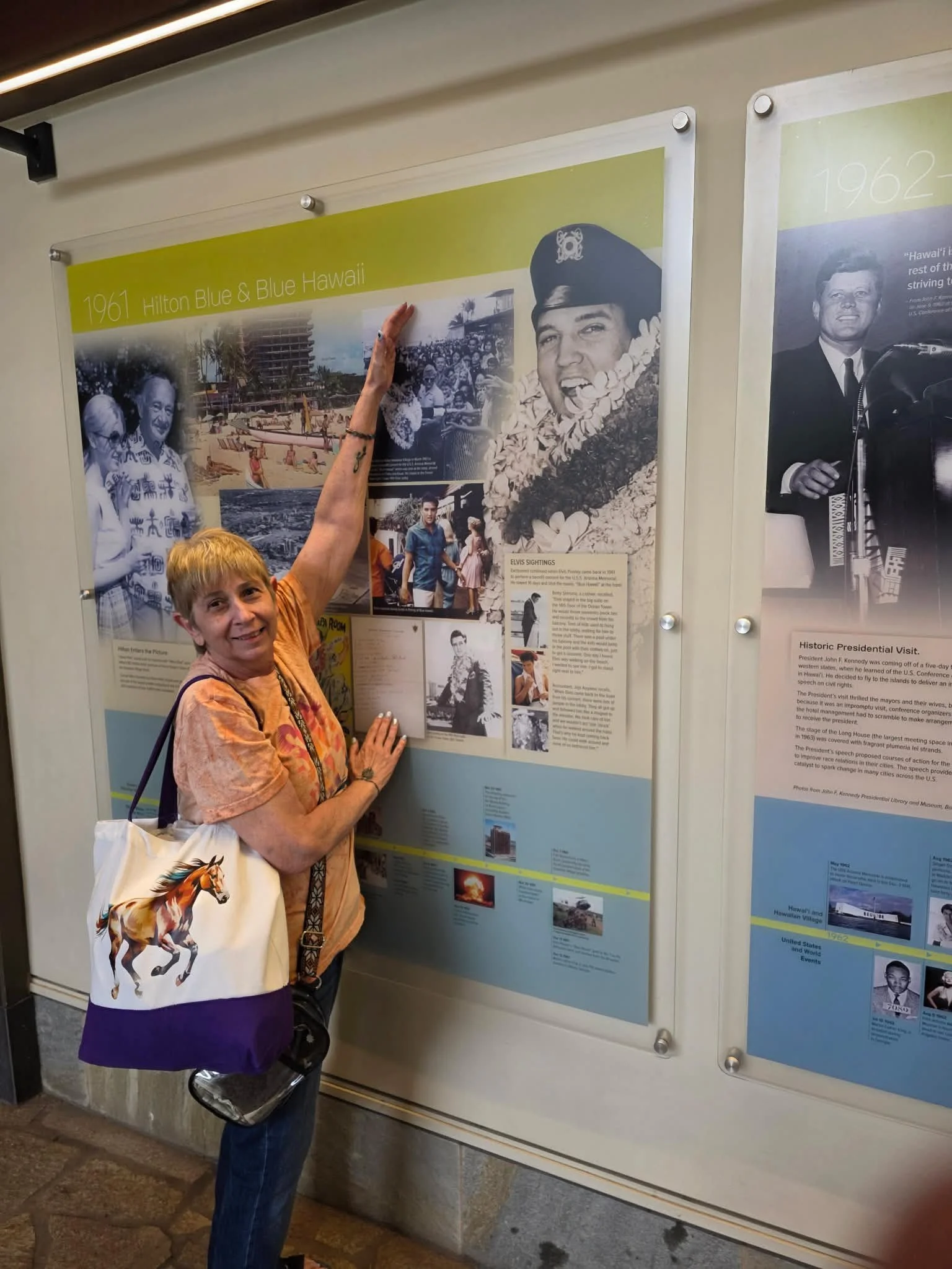 A smiling woman with short blond hair and a floral blouse standing next to an exhibit board at a museum, pointing at a photo of a young Elvis Presley wearing a lei and a cap, with other black and white photos of Elvis and historical information around.