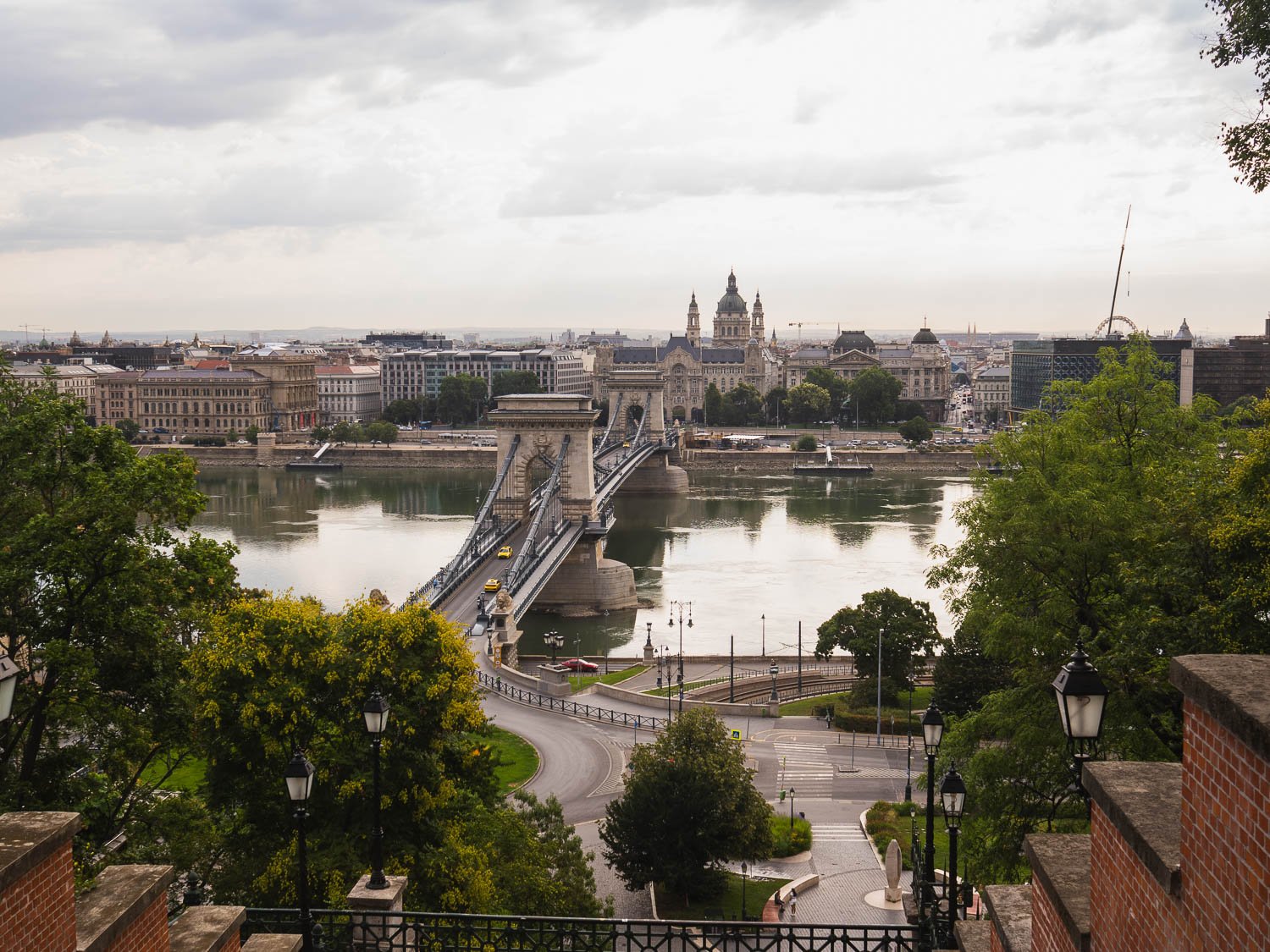 The Chain Bridge crossing the Danube, seen from the Buda side over trees and a brick parapet, with Pest and the dome of St Stephen's Basilica beyond