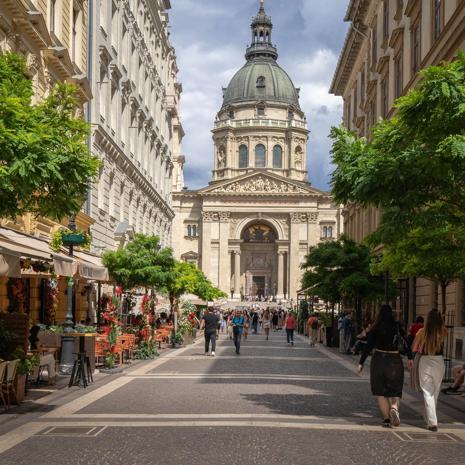 St Stephen's Basilica at the end of Zrínyi utca, a pedestrian street lined with trees and café terraces, people walking towards it