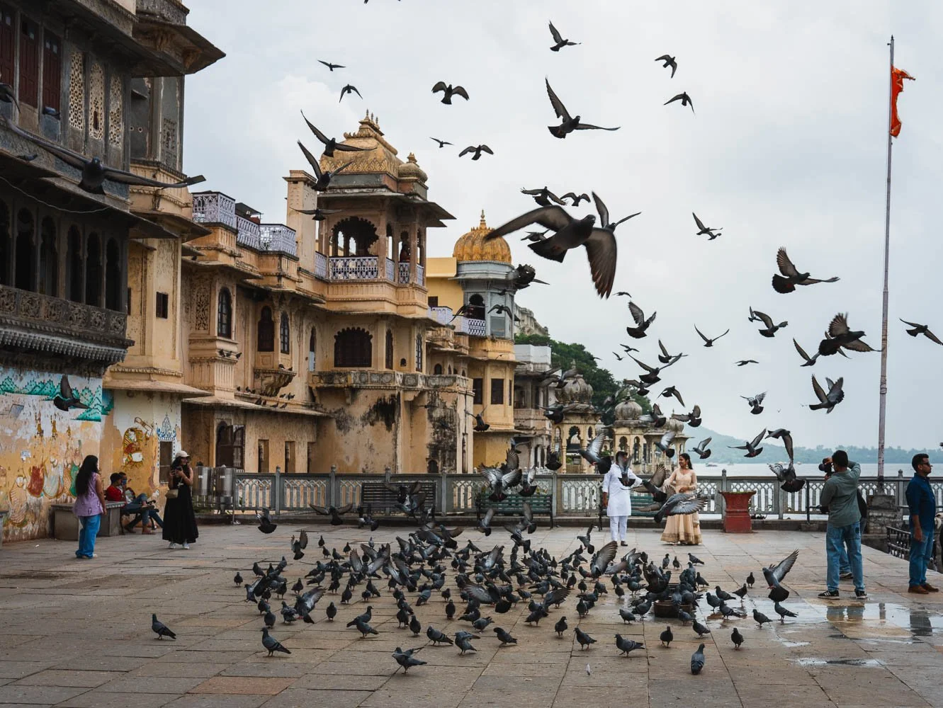 Pigeons rise over Gangaur Ghat beside Lake Pichola, a flagpole and old havelis behind