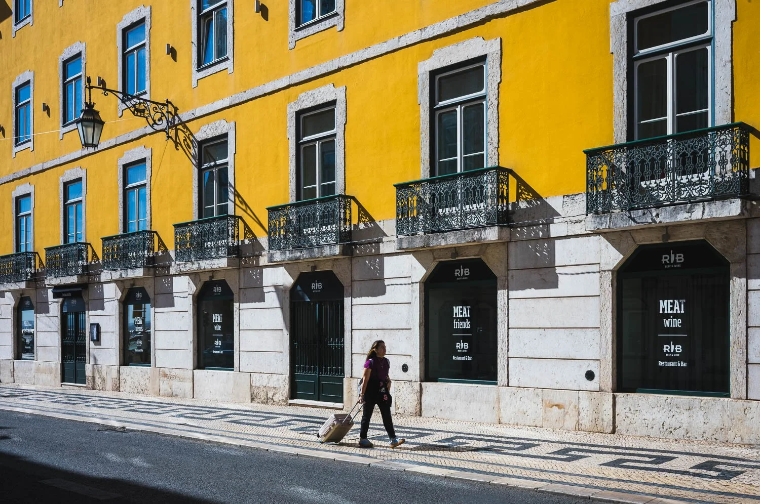 A woman walks with a suitcase past a yellow façade with wrought-iron balconies in the Baixa