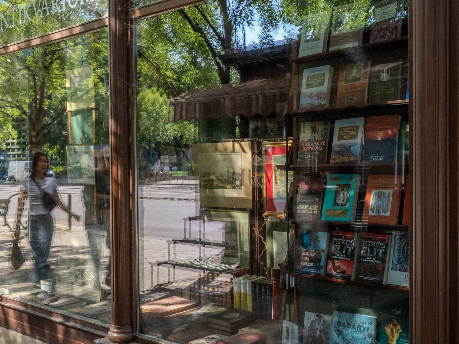 Window of a Budapest antiquarian bookshop full of old books and prints, a woman walking past reflected in the glass