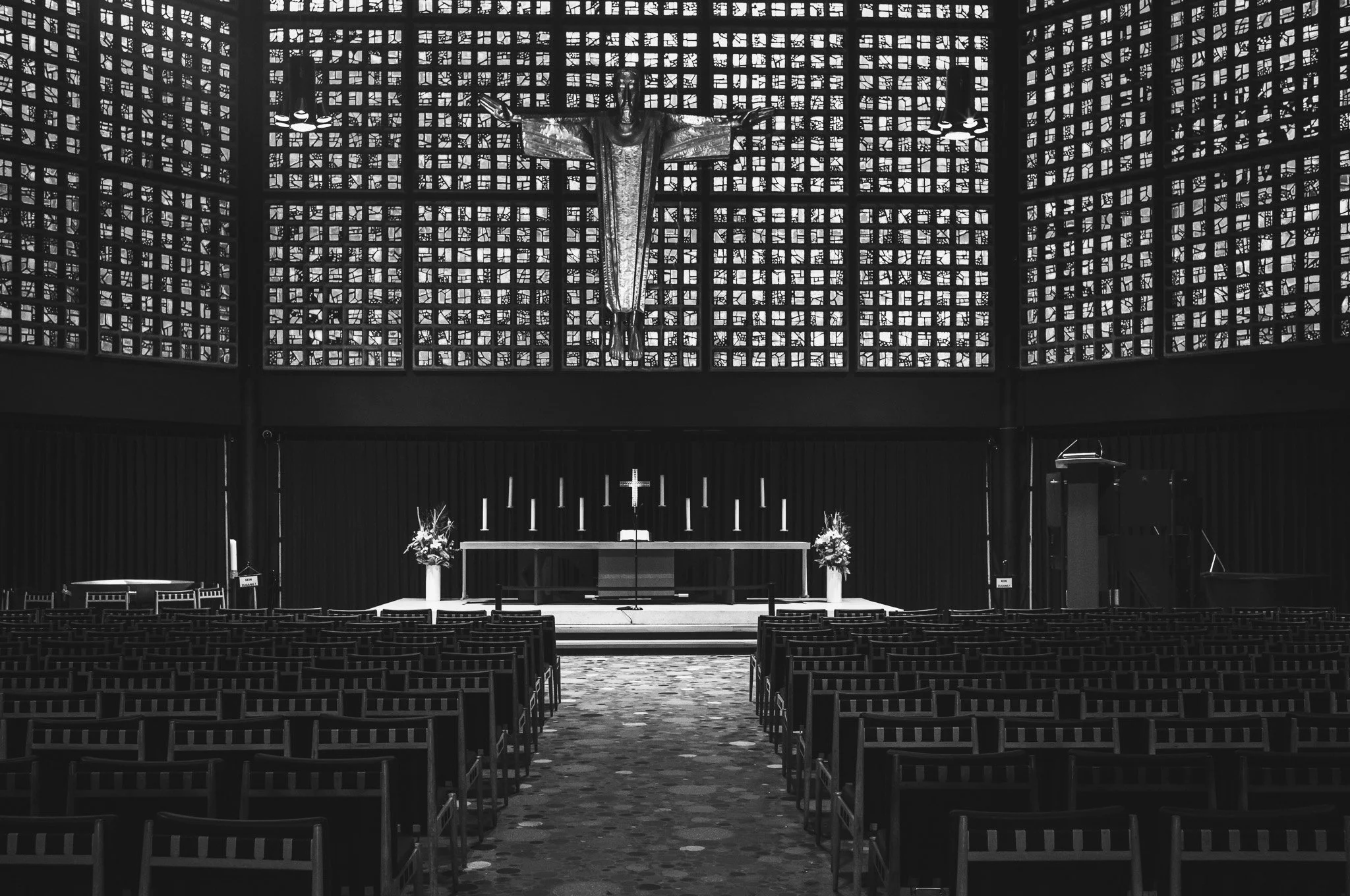 Black-and-white photograph of the interior of the Kaiser Wilhelm Memorial Church in Berlin, a figure of Christ above the altar against walls of glass blocks, rows of chairs below
