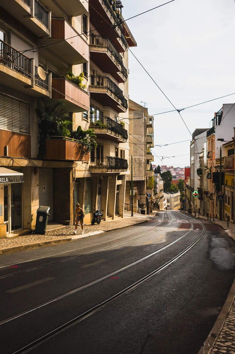 An empty Lisbon street curving between apartment buildings, tram lines and overhead wires in soft light
