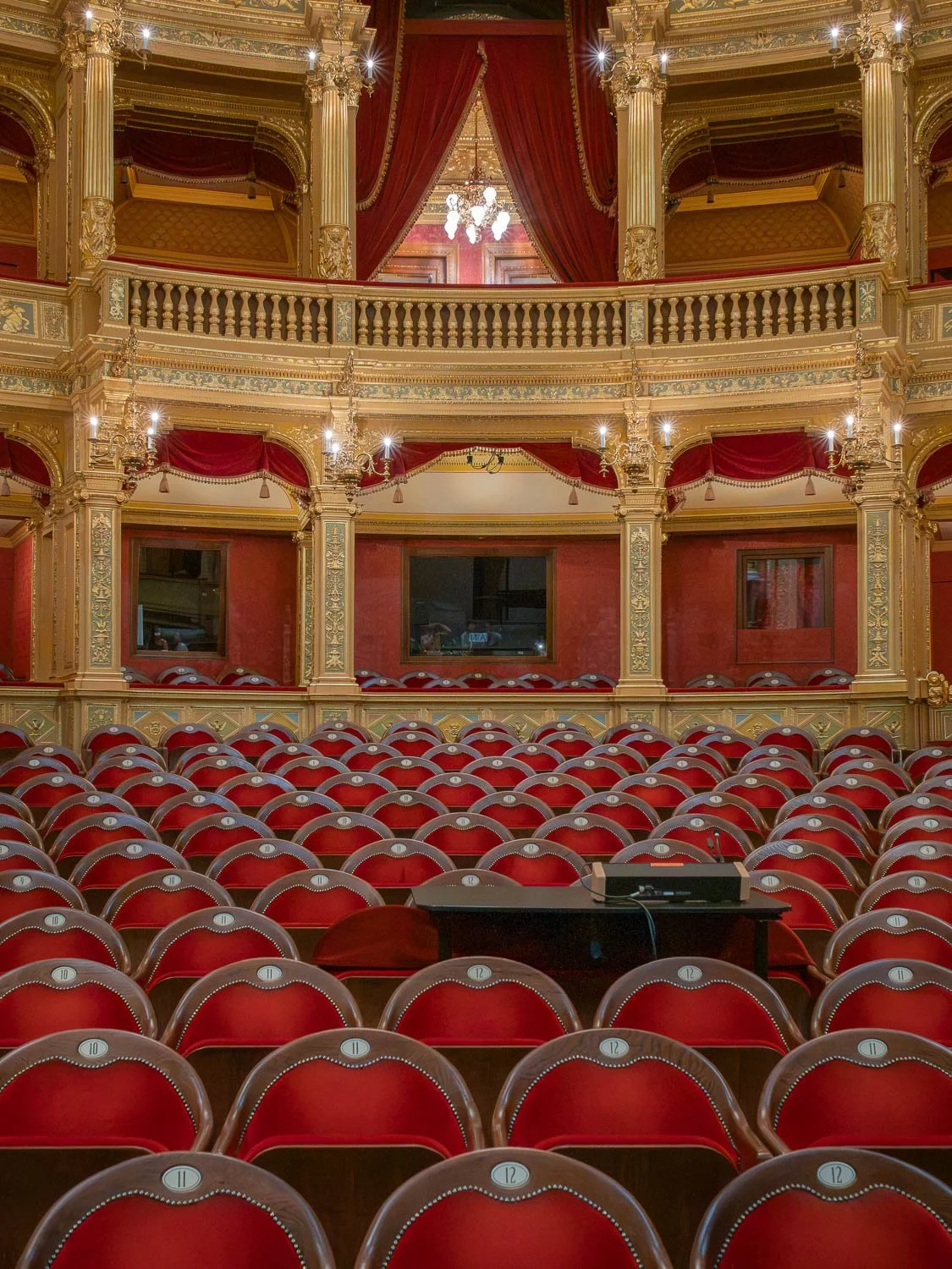 Rows of numbered red velvet seats facing tiers of gilded boxes with red curtains in an ornate Budapest theatre auditorium