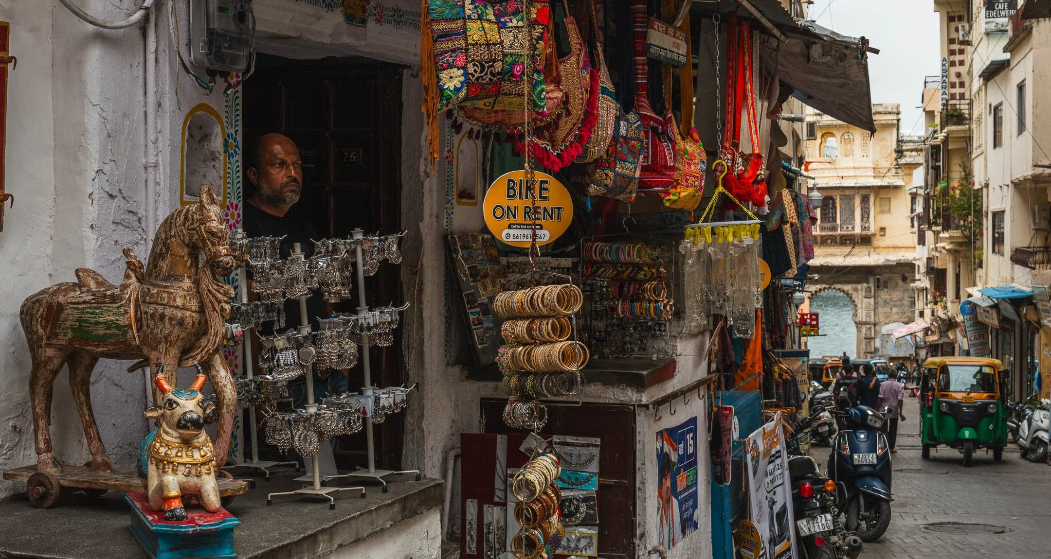 A shopkeeper watches from his doorway beside a painted wooden horse, the lane behind leading to an arch over the lake