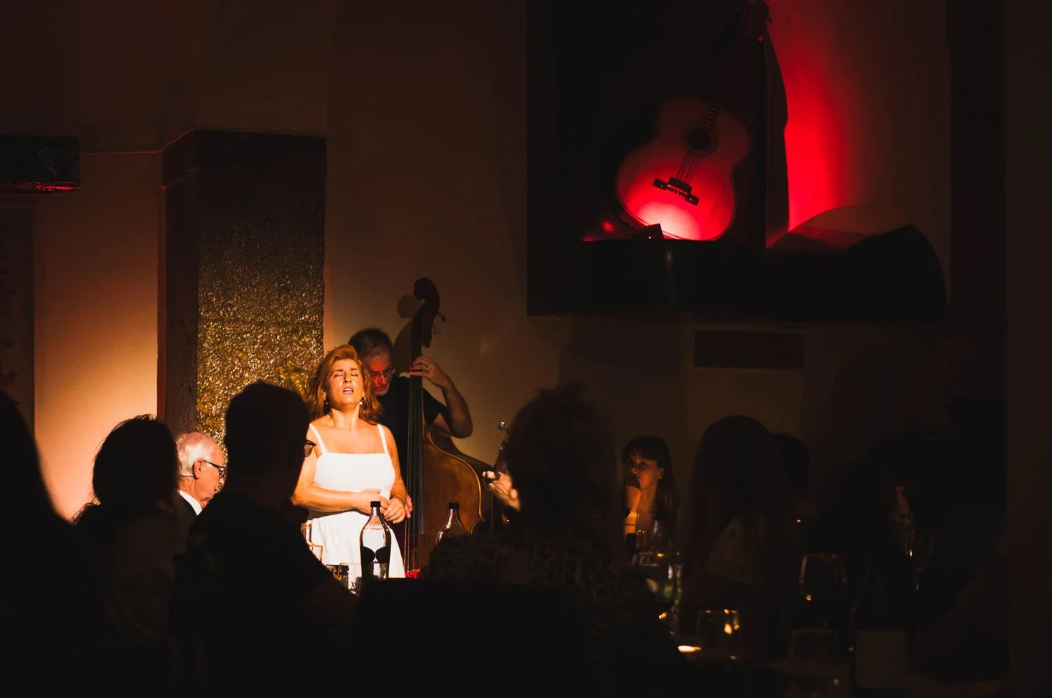 A fado singer in a white dress performs in a dim, red-lit restaurant, the audience in silhouette