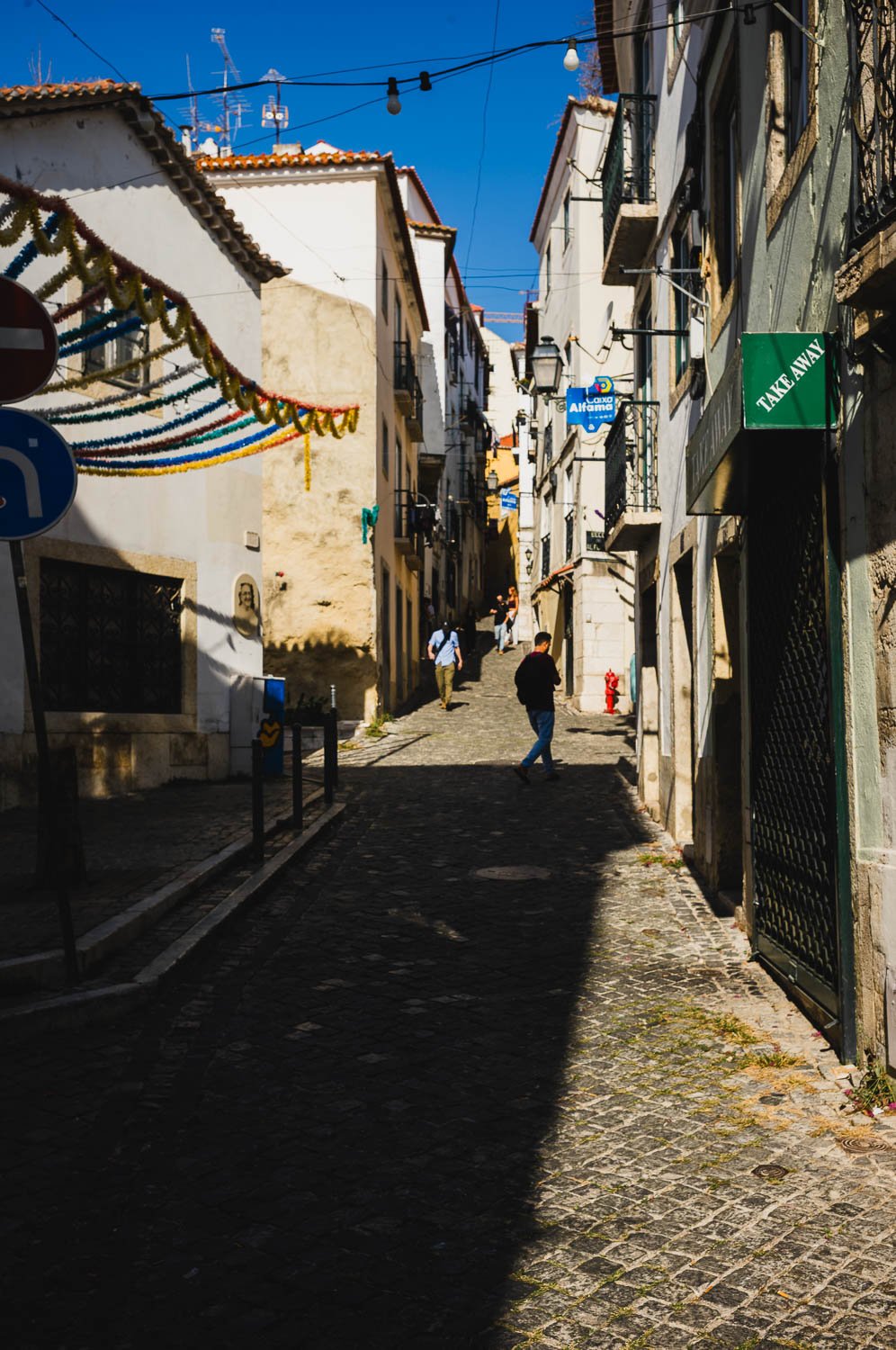 A steep, shaded alley in old Lisbon strung with festival bunting, a figure walking at the far end