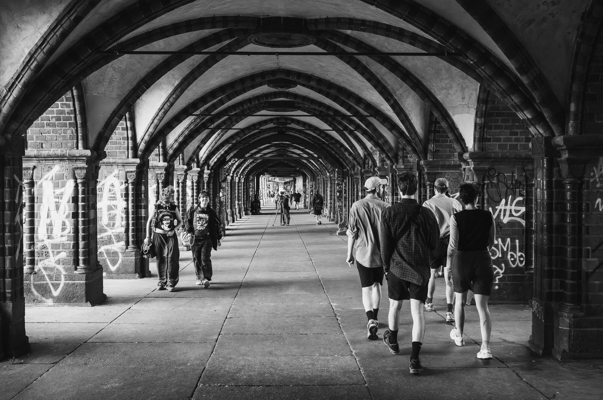 Black-and-white photograph of people walking through the vaulted brick arcade beneath the Oberbaum Bridge in Berlin, its pillars marked with graffiti