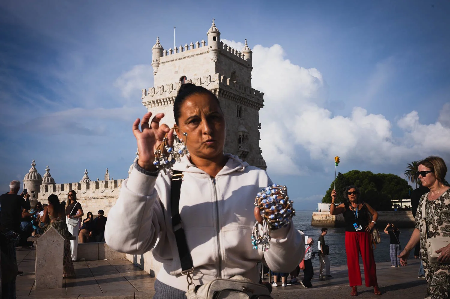 A street vendor holds up beaded bracelets in front of the Belém Tower, tourists behind her