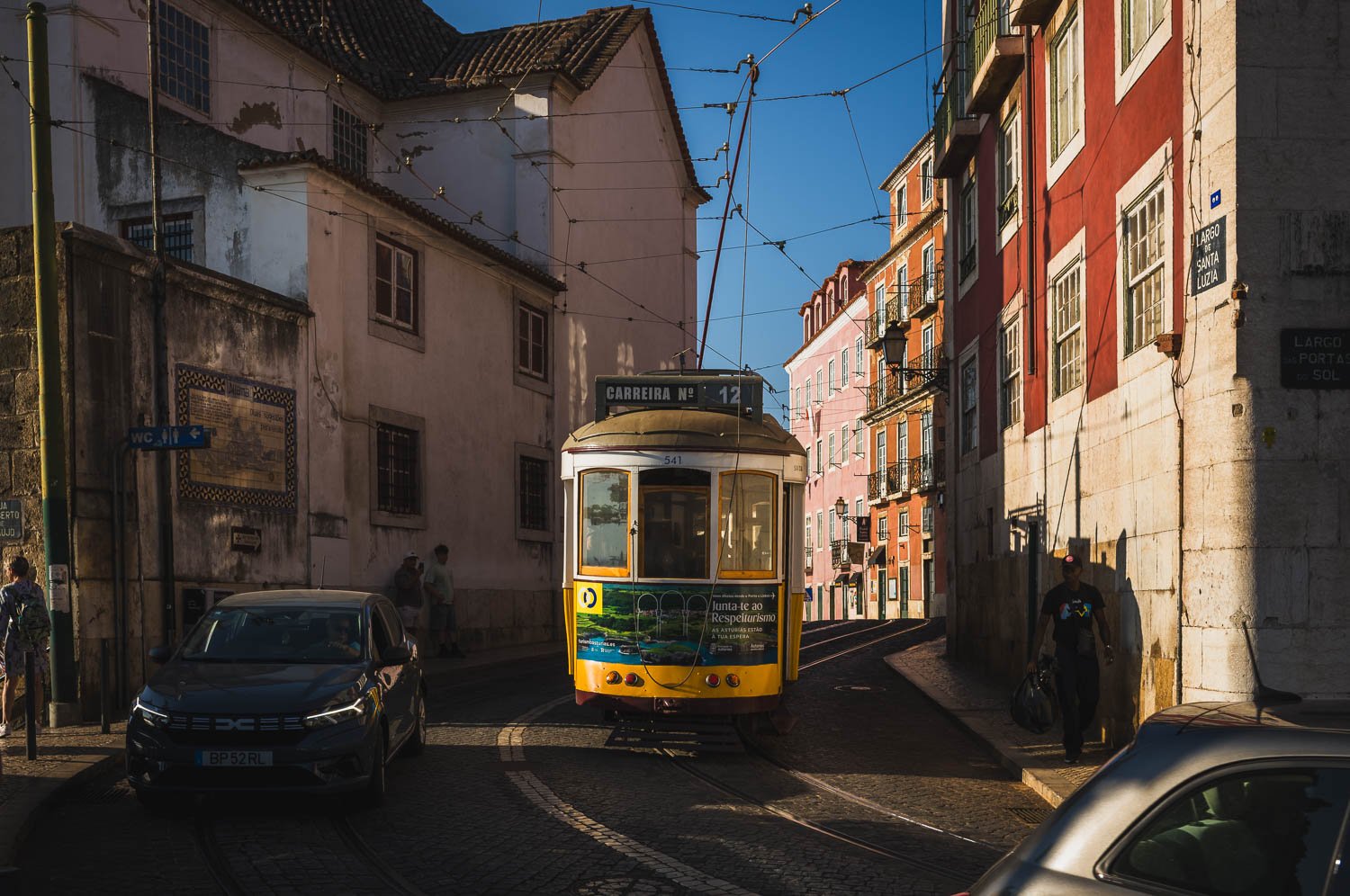 A yellow tram climbs a narrow street between old houses, a parked car in the foreground