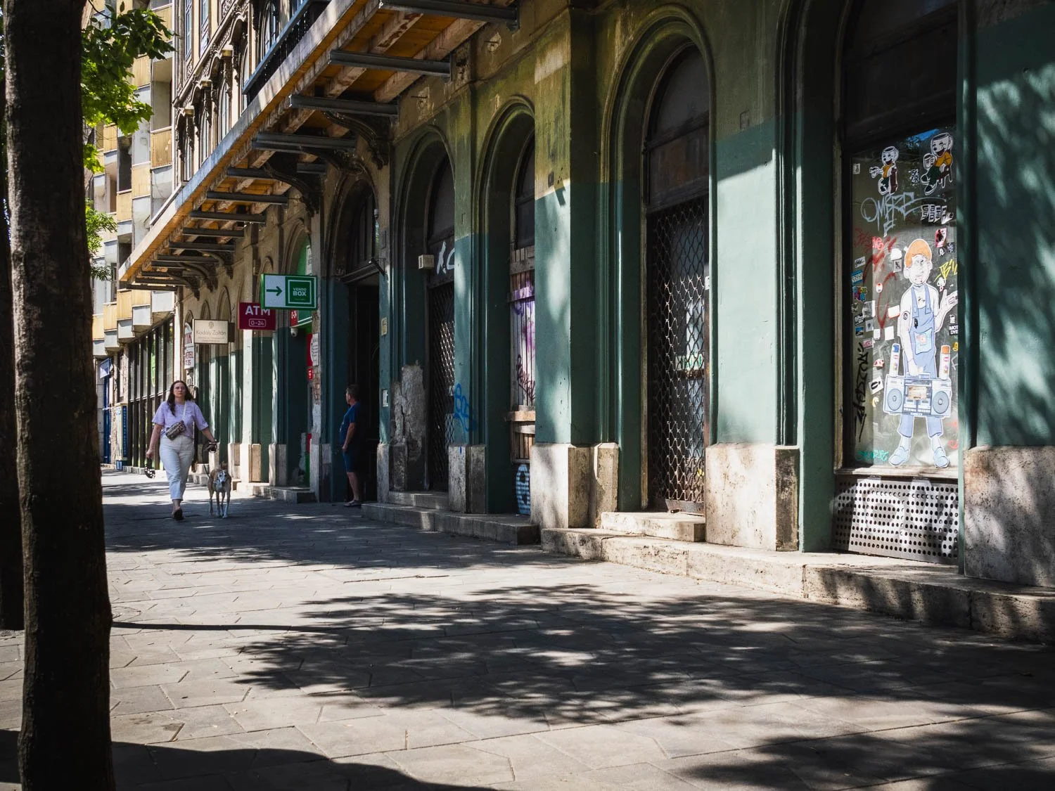 A woman walks her dog along a shaded arcade of green arches and shuttered shopfronts on a Budapest street