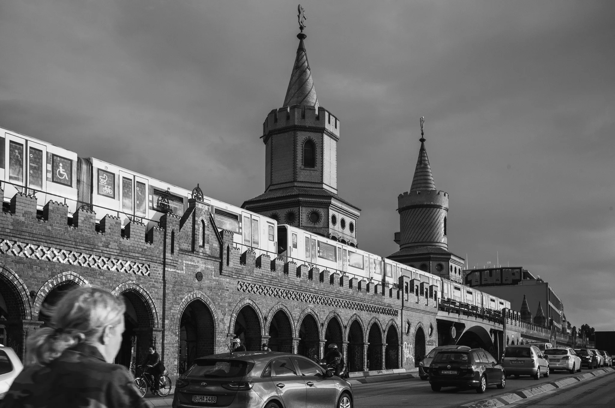 Black-and-white photograph of a U-Bahn train crossing the Oberbaum Bridge in Berlin, its brick towers and arches behind a woman in the foreground