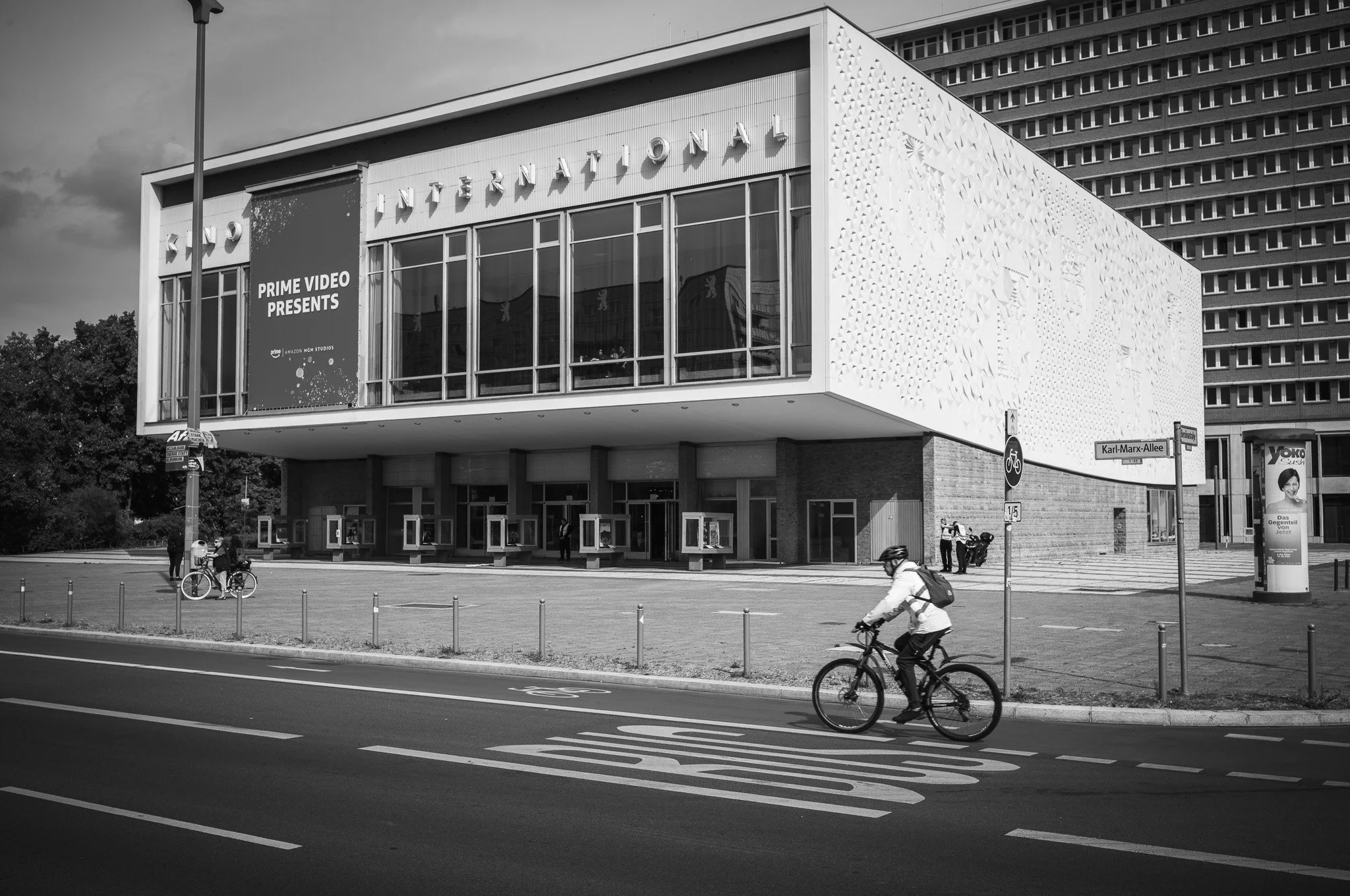 Black-and-white photograph of the Kino International cinema on Karl-Marx-Allee, Berlin, with a cyclist riding along the bus lane in front