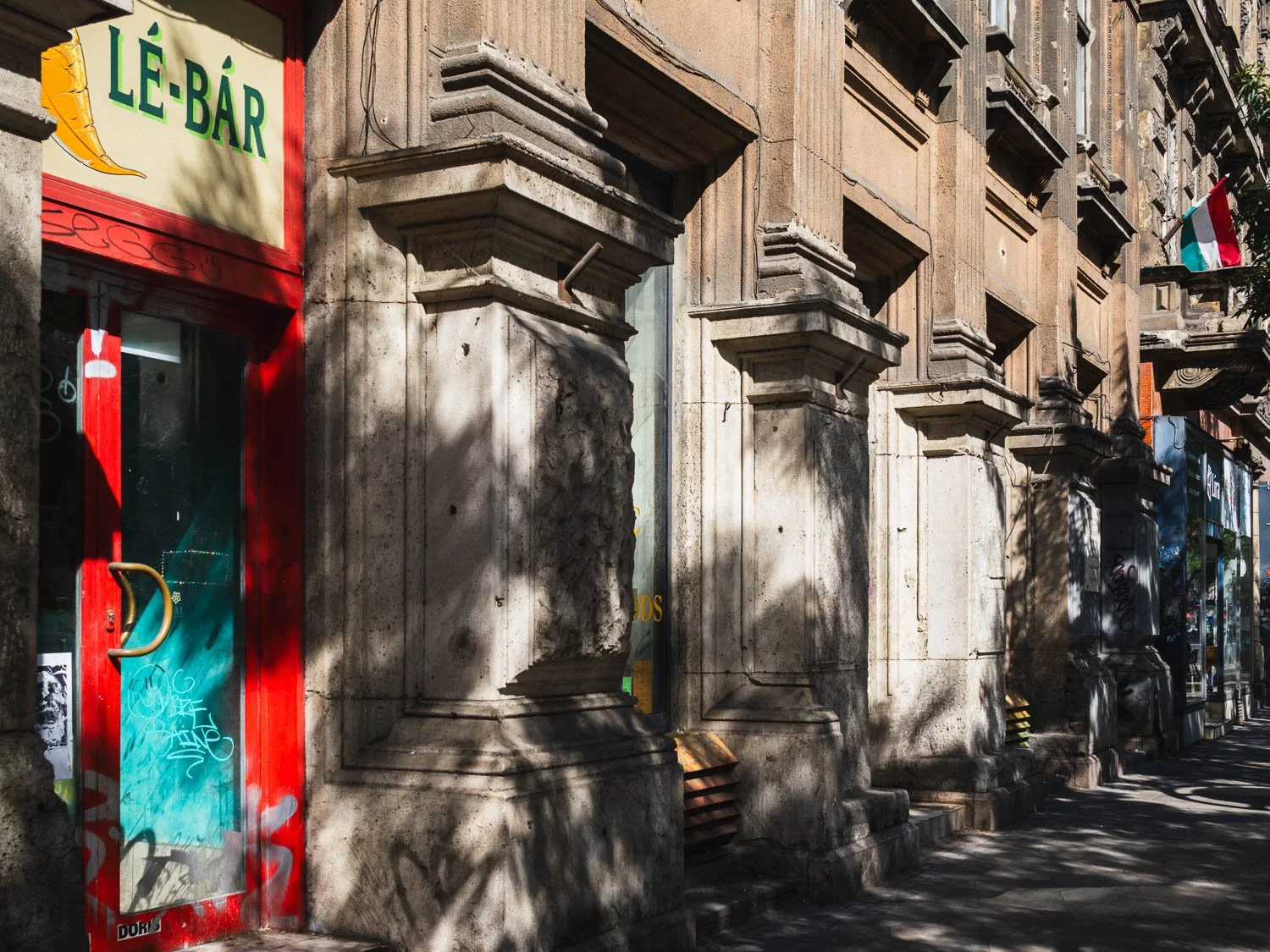 Red-framed door of the Lé-Bár juice bar beside a weathered stone façade in dappled shade, a Hungarian flag on a balcony above
