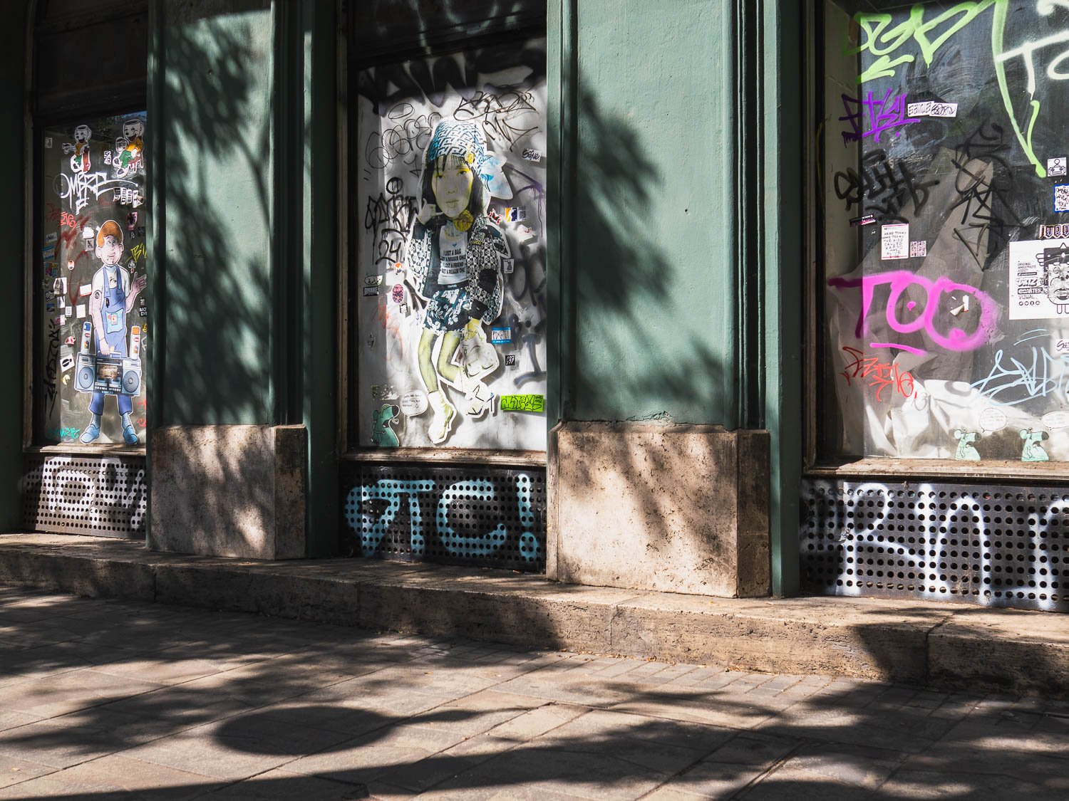 Green-painted shopfronts covered in posters, stickers and graffiti, in dappled tree shade on a Budapest pavement