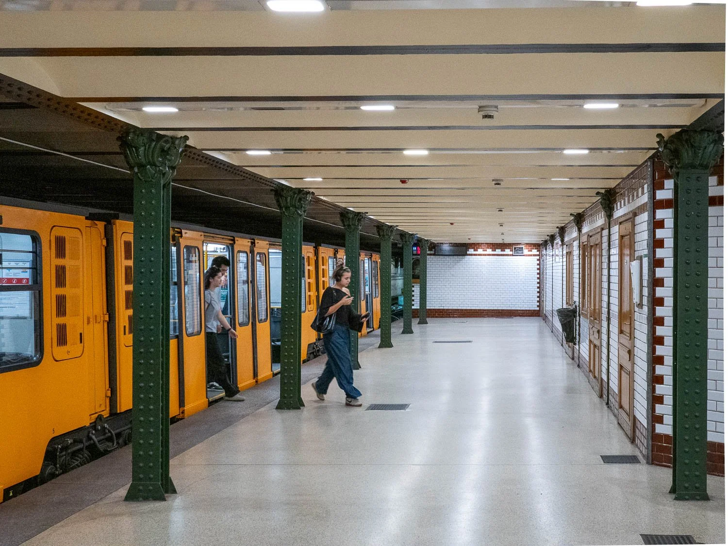 Platform of the Millennium Underground with green cast-iron columns, white tiled walls and a yellow train, a woman stepping off while looking at her phone