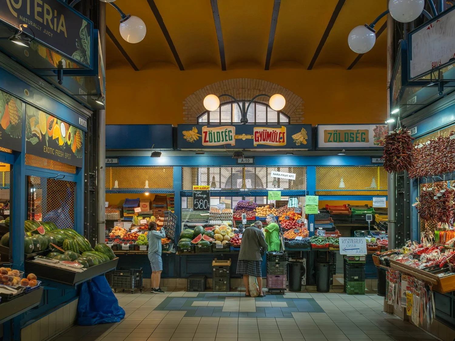 Fruit and vegetable stall inside a Budapest market hall, shoppers at the counter and strings of dried red paprika hanging at the right