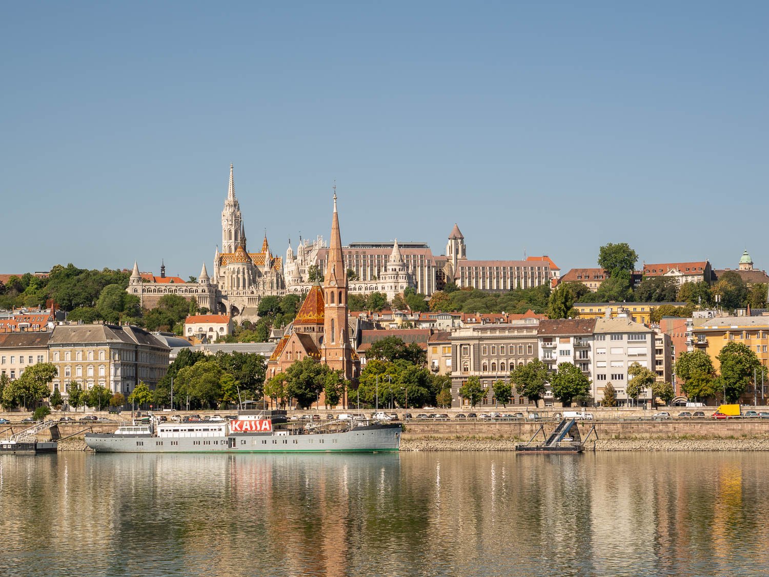 Buda riverfront with Matthias Church and the Fishermen's Bastion on Castle Hill, the red-brick spire of the Szilágyi Dezső tér Reformed Church below, and a boat moored on the Danube