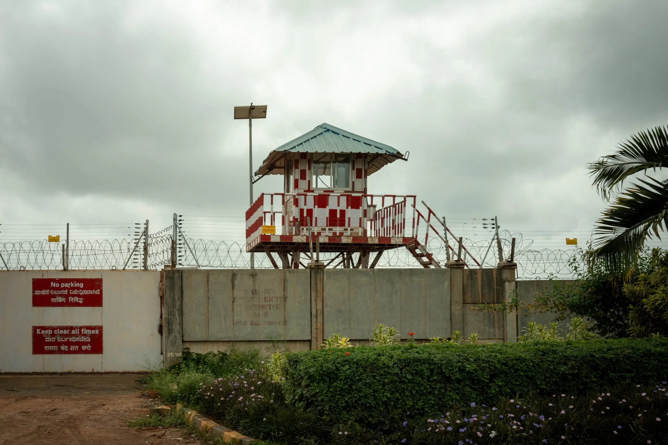A red-and-white watchtower above a perimeter wall topped with razor wire, under heavy cloud