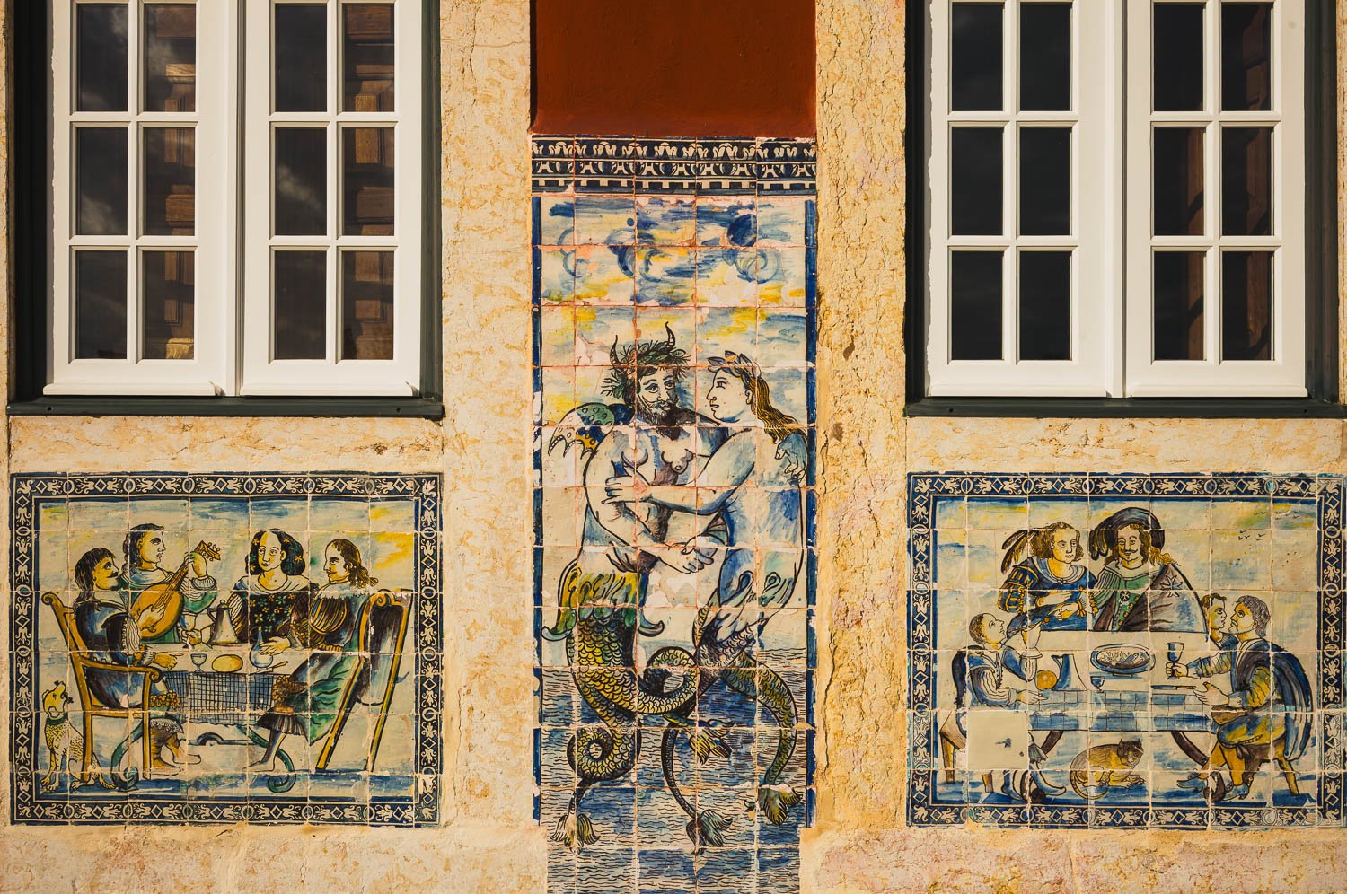 Old azulejo panels of figures and a mermaid between white-framed windows on an ochre wall