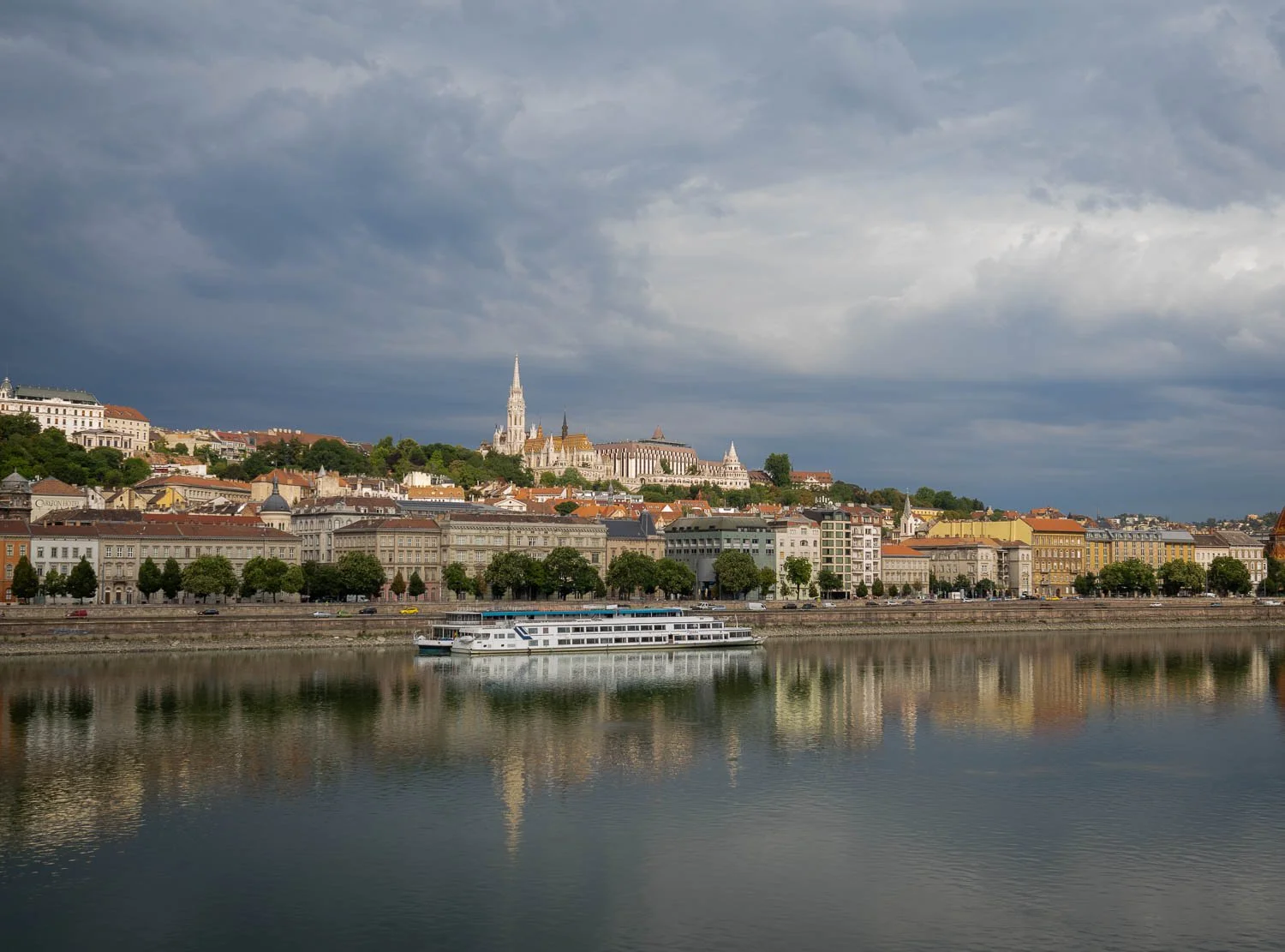 Buda riverfront and Castle Hill with the spire of Matthias Church, seen across a still Danube under heavy grey clouds, a river cruiser moored below