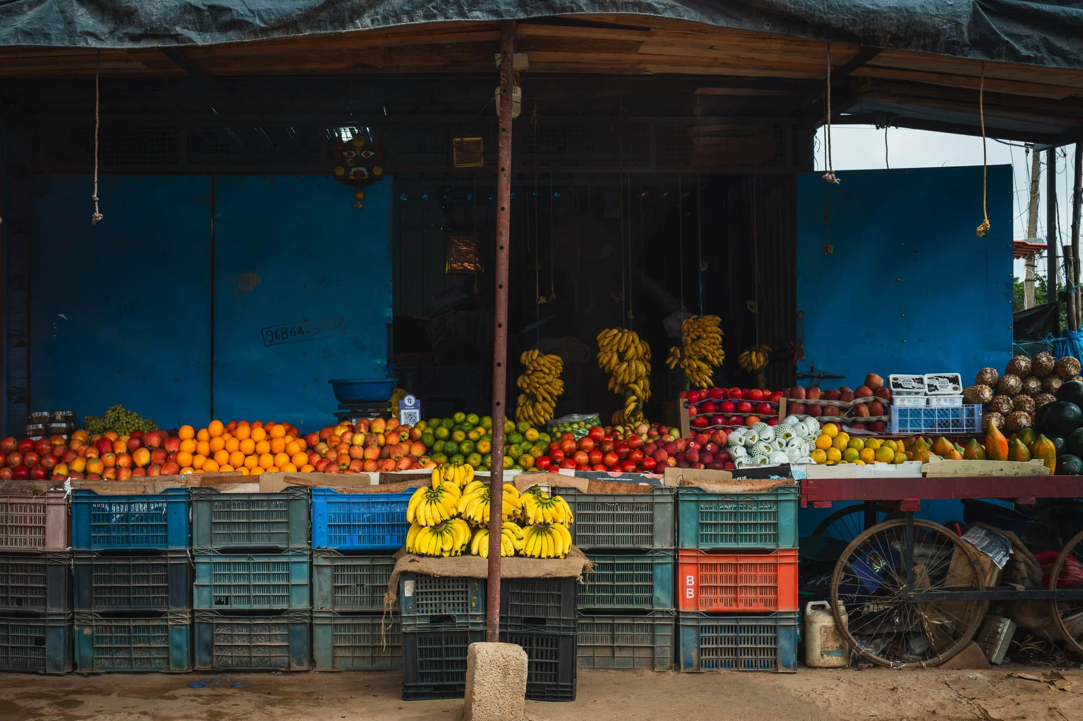 A roadside fruit stall stacked with apples, oranges and bananas on plastic crates, a cart wheel at its side