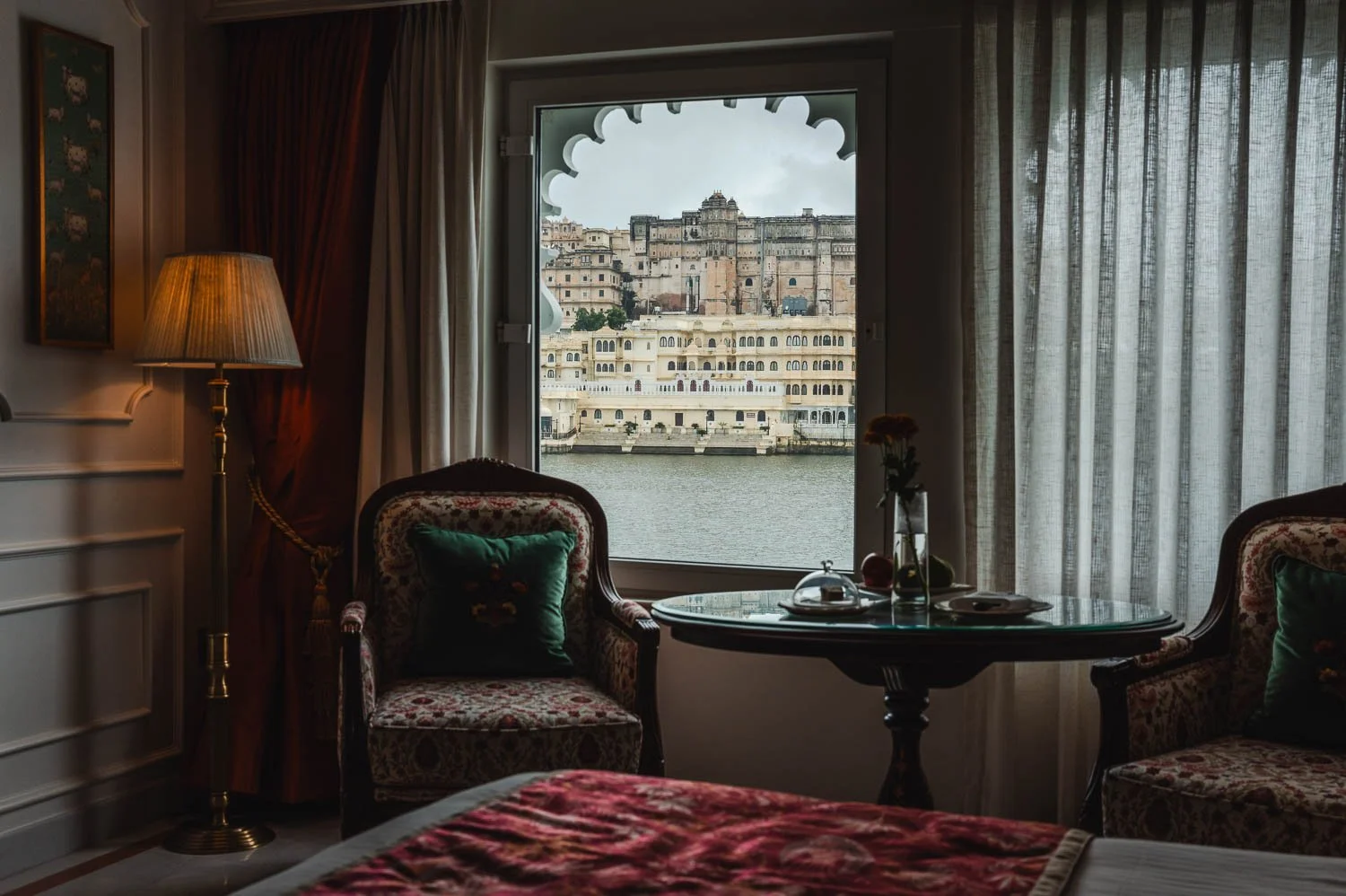 A hotel room with armchairs and a lamp, its scalloped window framing the City Palace across the lake
