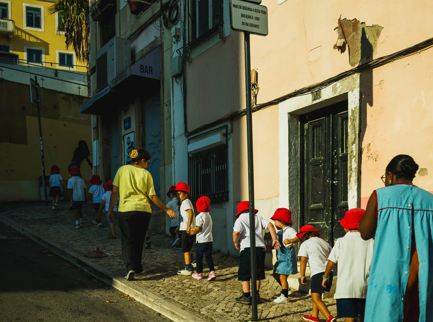A line of schoolchildren in red caps walks with their teachers up a narrow, sunlit Lisbon street