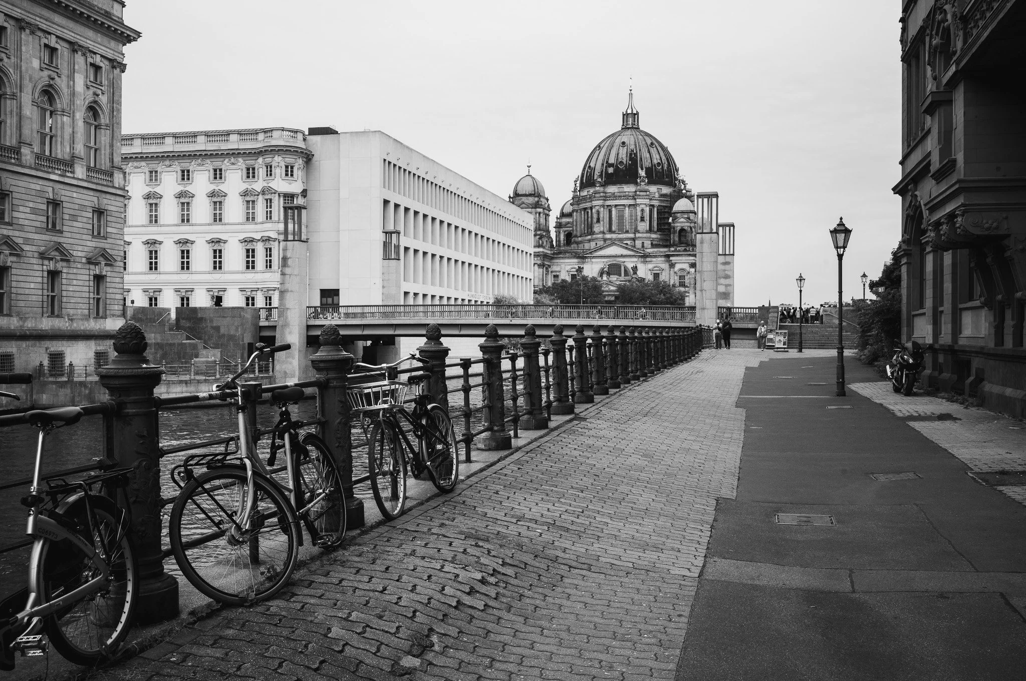 Black-and-white photograph of a riverside promenade with bicycles against iron railings, the Humboldt Forum and the dome of Berlin Cathedral beyond