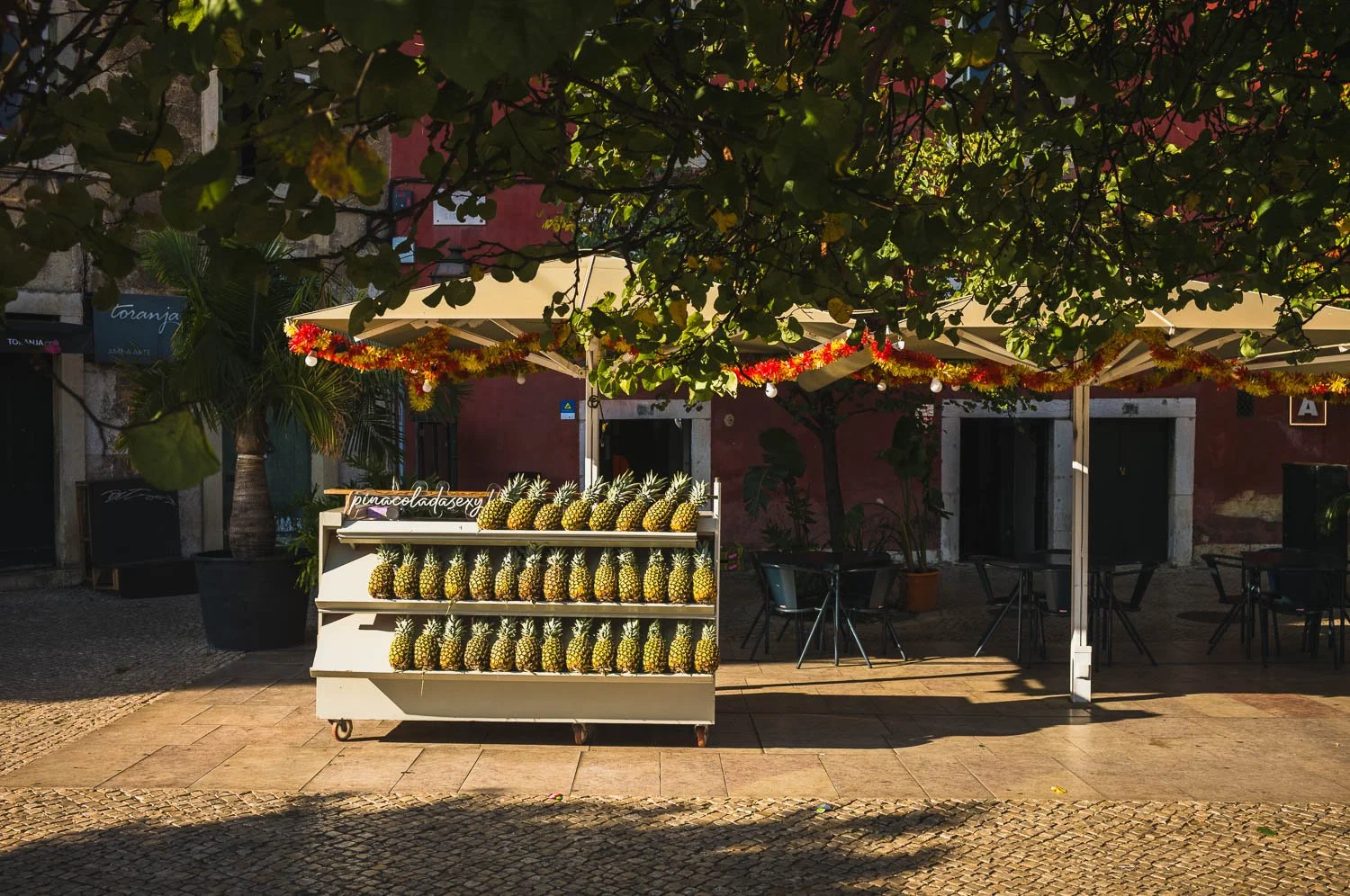 A cart stacked with pineapples stands under trees beside café umbrellas in a Lisbon square