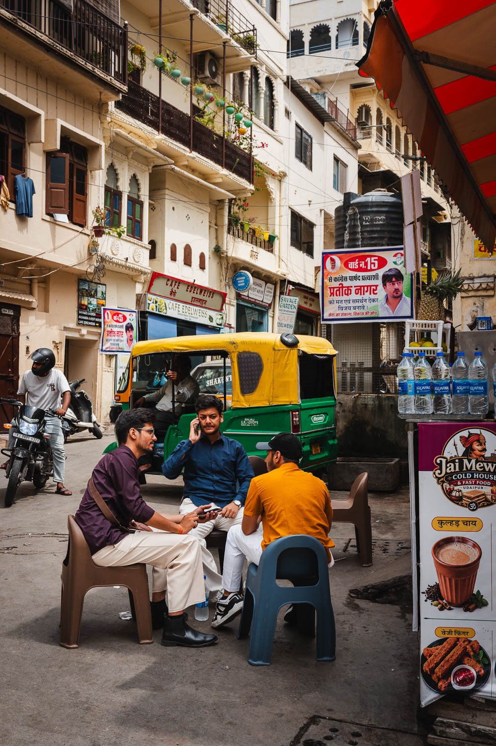 Men drink tea on plastic chairs at a chai stall, a yellow-and-green autorickshaw behind