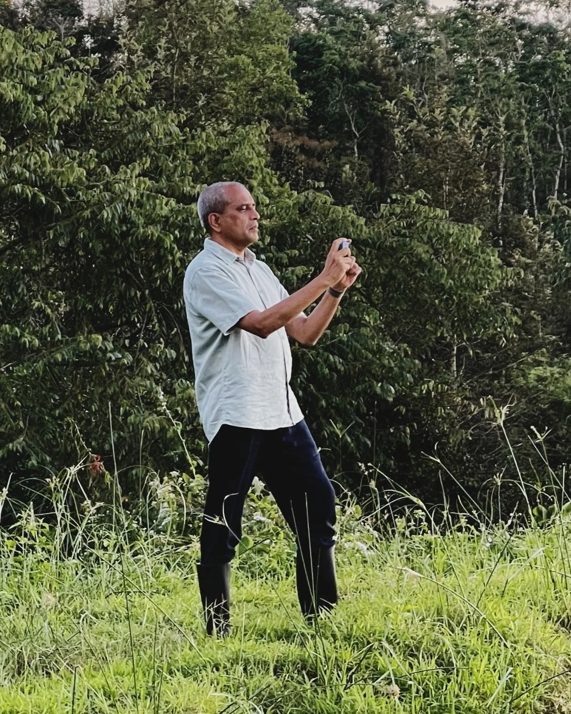 Shashikiran Mullur stands in tall grass at the edge of a forest, taking a photograph with his phone