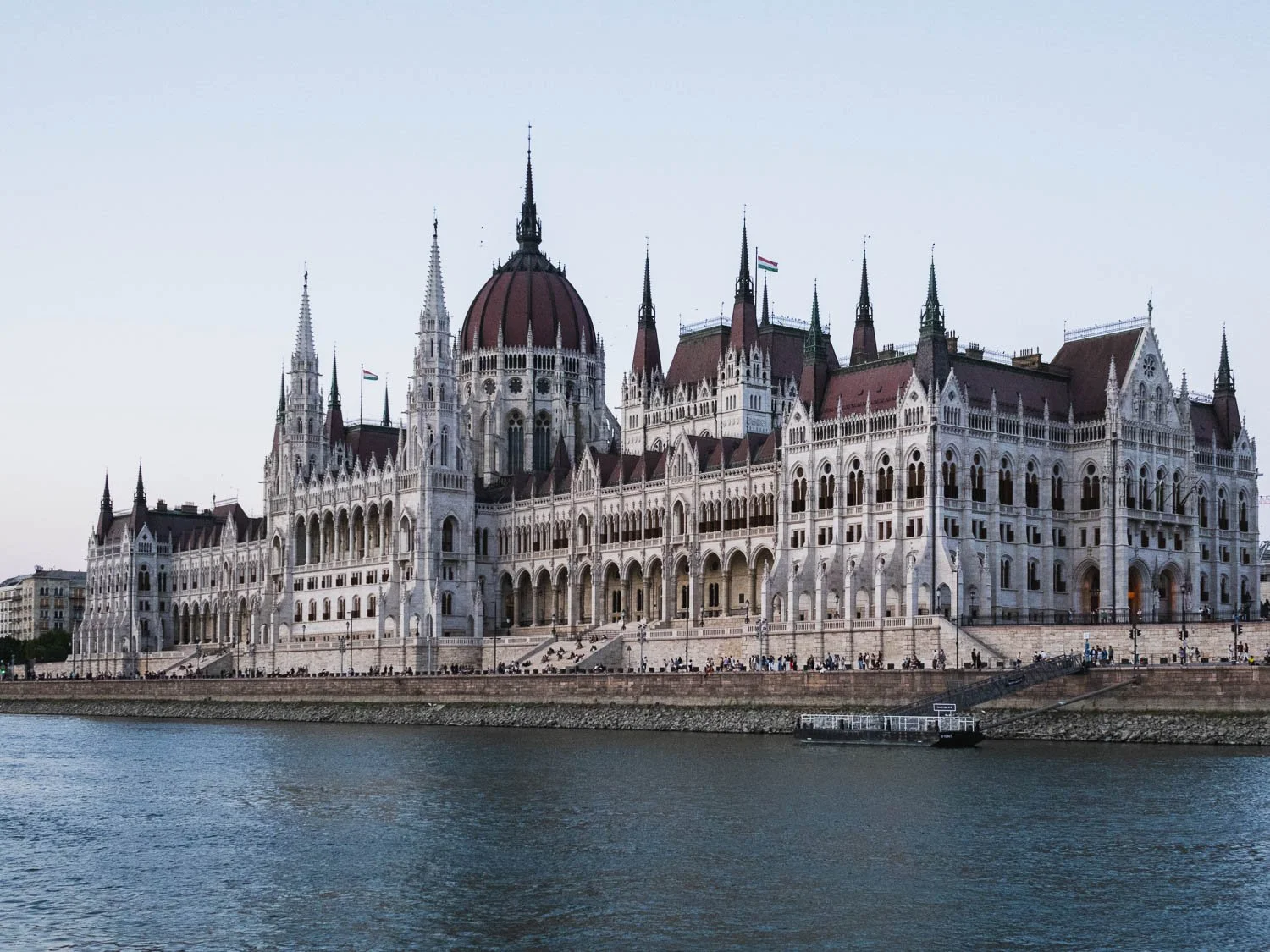 The Hungarian Parliament Building on the bank of the Danube, its central dome and Gothic Revival spires against a pale evening sky