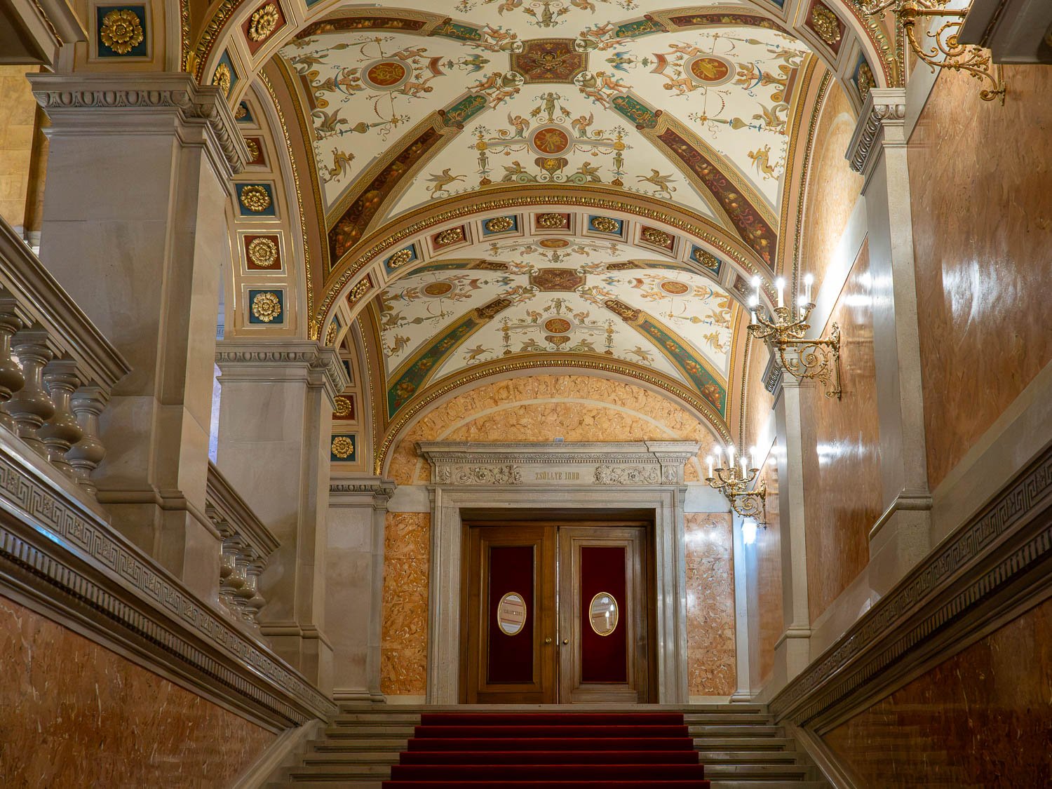 Grand staircase with marble walls and a red carpet rising under a painted and gilded vaulted ceiling to a pair of wooden doors