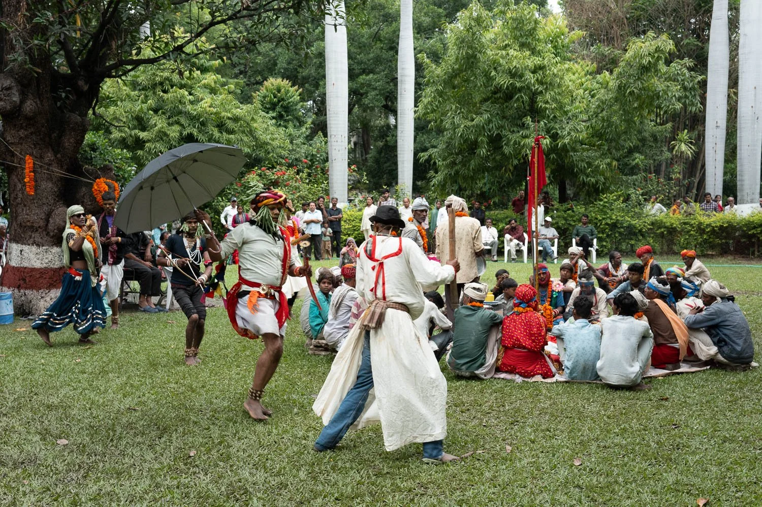 Folk dancers perform on a lawn before a seated crowd, one twirling an umbrella