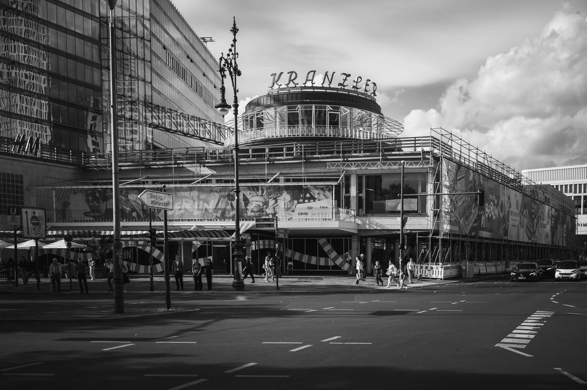 Black-and-white photograph of the Café Kranzler rotunda on Kurfürstendamm in Berlin, partly wrapped in scaffolding, people crossing the street below