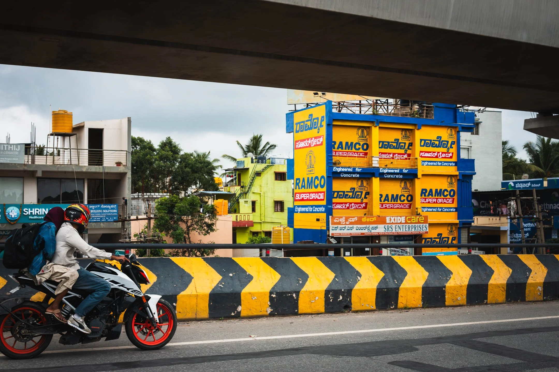 Two riders on a motorbike pass under a flyover, a Ramco cement billboard and painted buildings beyond