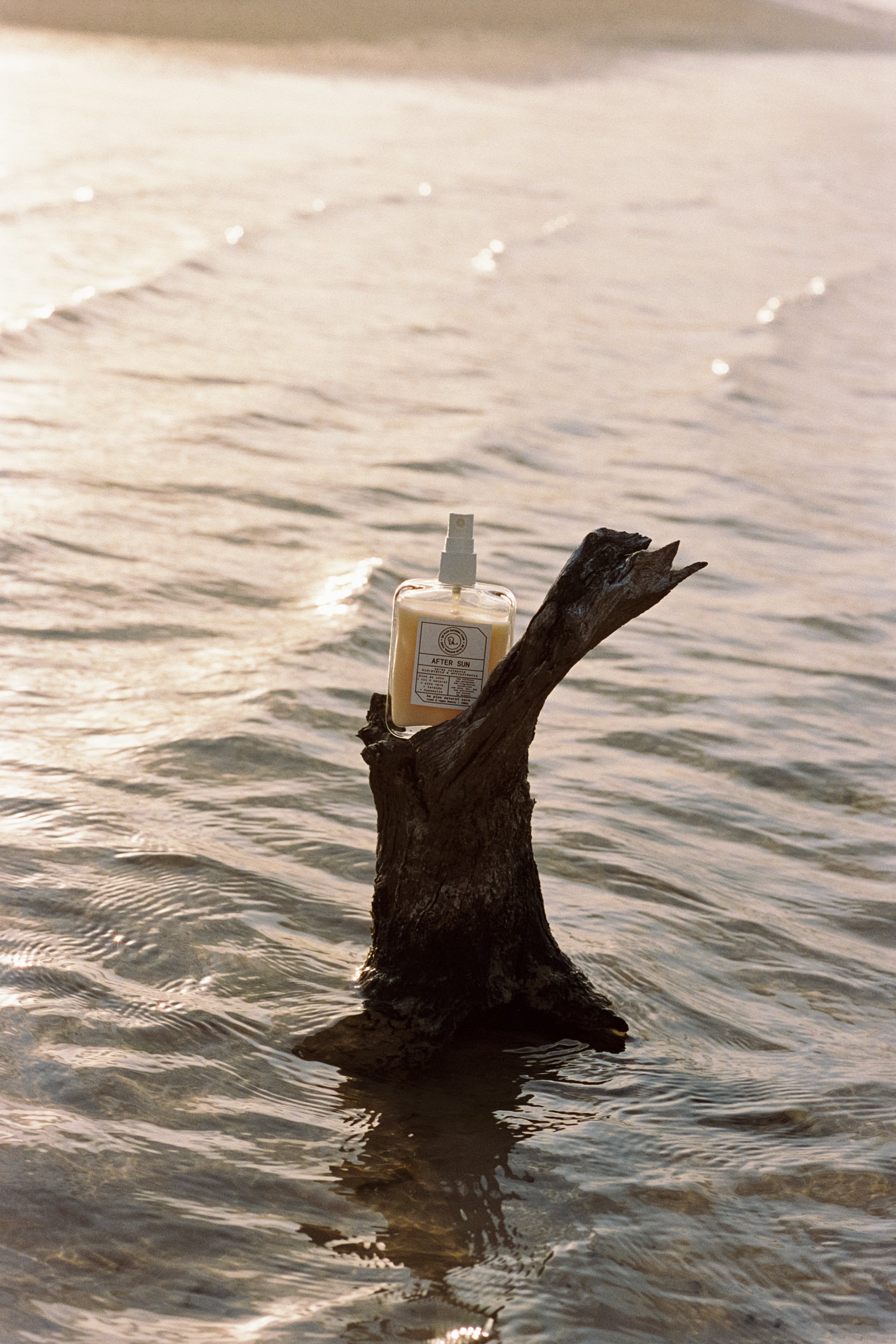 After Sun spray bottle balanced on a driftwood stump in rippling water in lagoon, sunset, Be Plus Natural Care, Lençóis Maranhenses, Brazil, on 35mm film.