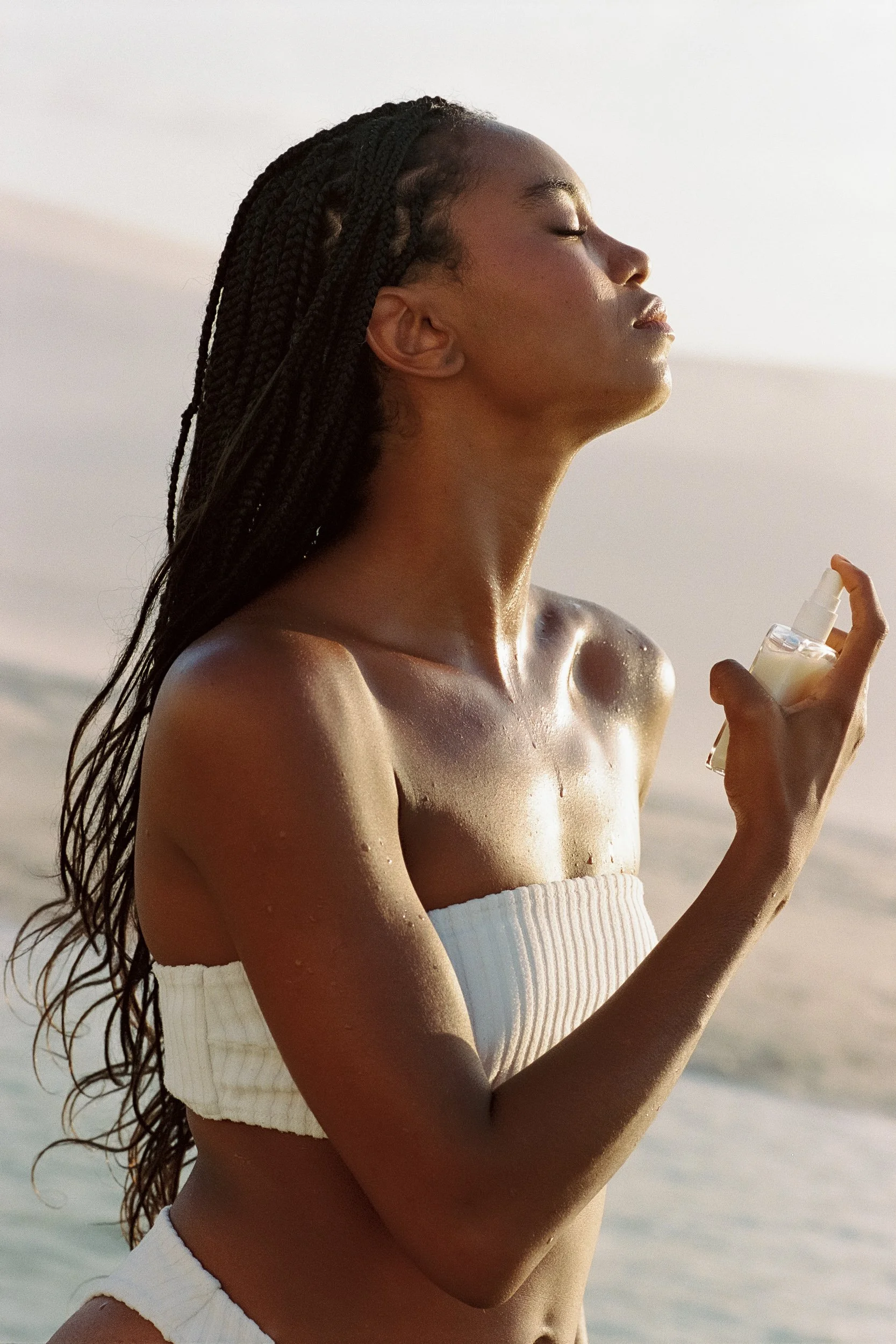 Model in a white bandeau top misting her neck with eyes closed and sunlit skin, Be Plus Natural Care, Lençóis Maranhenses, Brazil, on 35mm film.