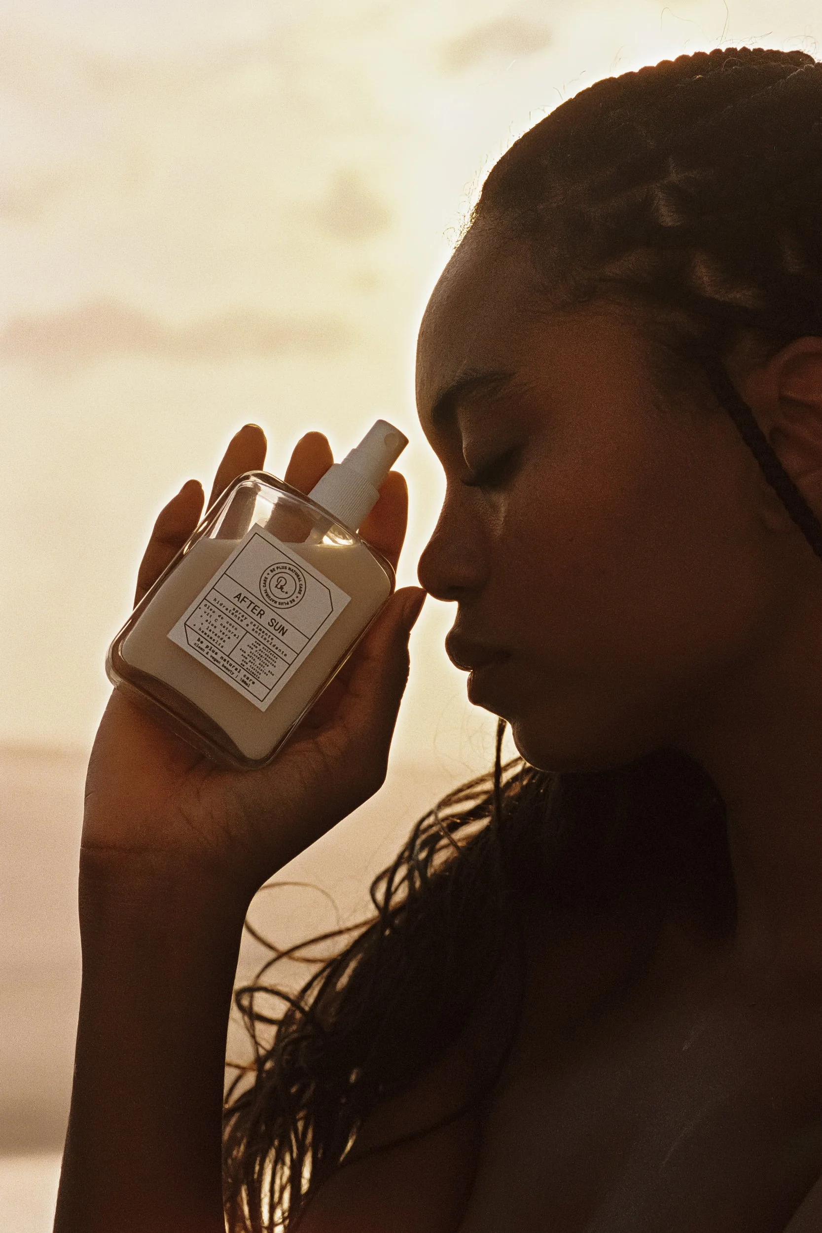 Backlit profile of a model with eyes closed holding a product After Sun spray at sunset, Be Plus Natural Care, Lençóis Maranhenses, Brazil, on 35mm film.