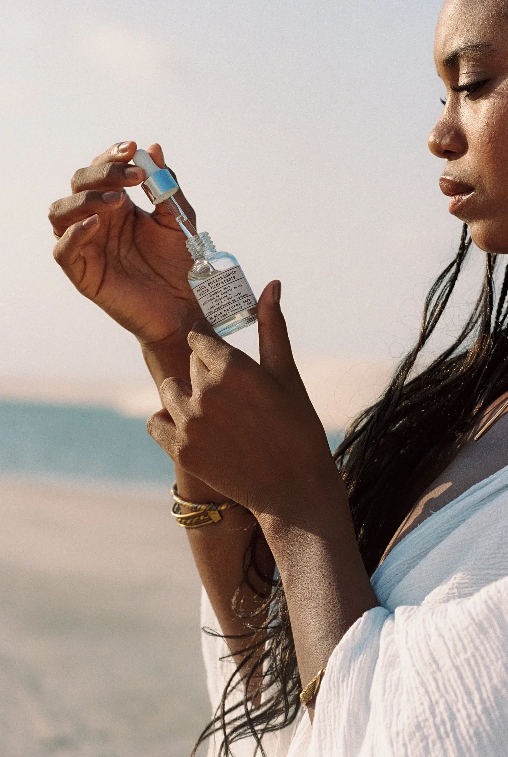 Close-up of hands lifting the dropper from a clear glass multi antioxidant serum bottle, Be Plus Natural Care, Lençóis Maranhenses, Brazil, on 35mm film.