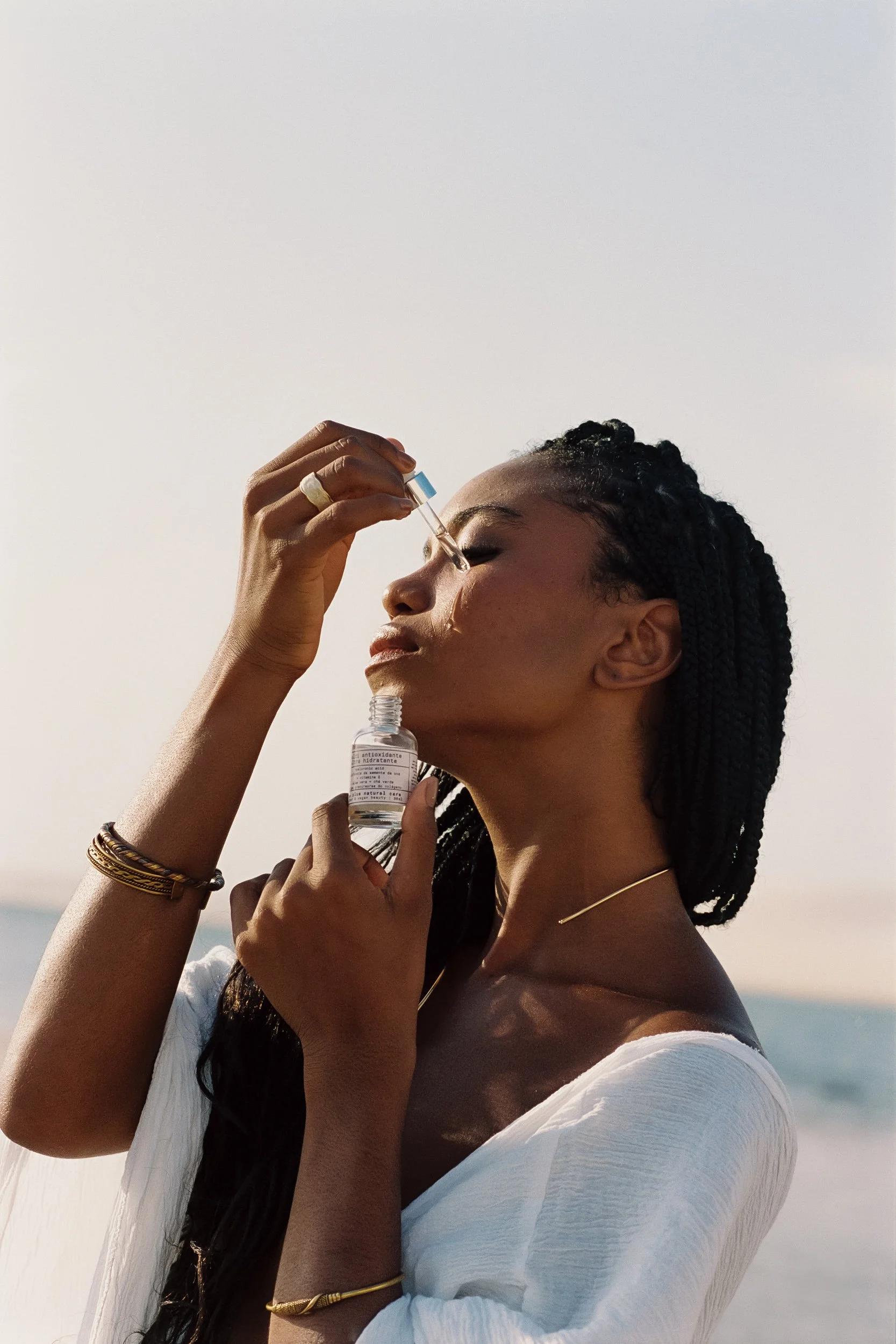 Model with head tilted back applying serum under her face with a dropper, Be Plus Natural Care, at golden hour, Lençóis Maranhenses, Brazil, on 35mm film.