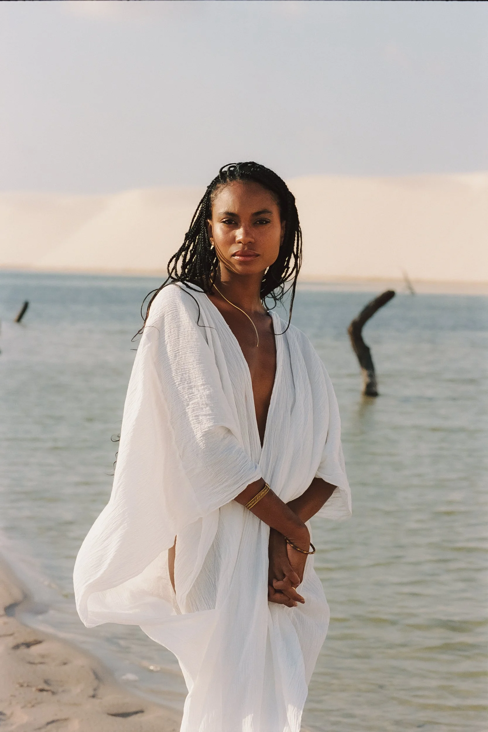Portrait of a model in a draped white kaftan by a lagoon dotted with driftwood, Be Plus Natural Care, at golden hour, Lençóis Maranhenses, Brazil, on 35mm film.